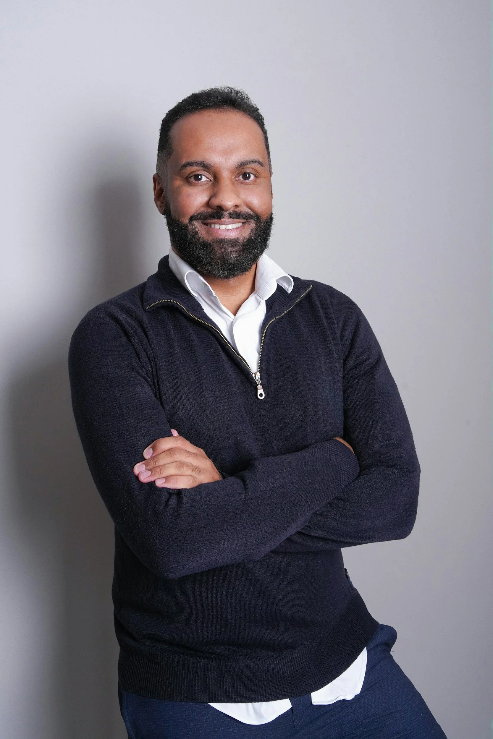 A man with dark hair and a beard smiling, crossing his arms, wearing a navy sweater over a white shirt, standing against a plain light gray wall.