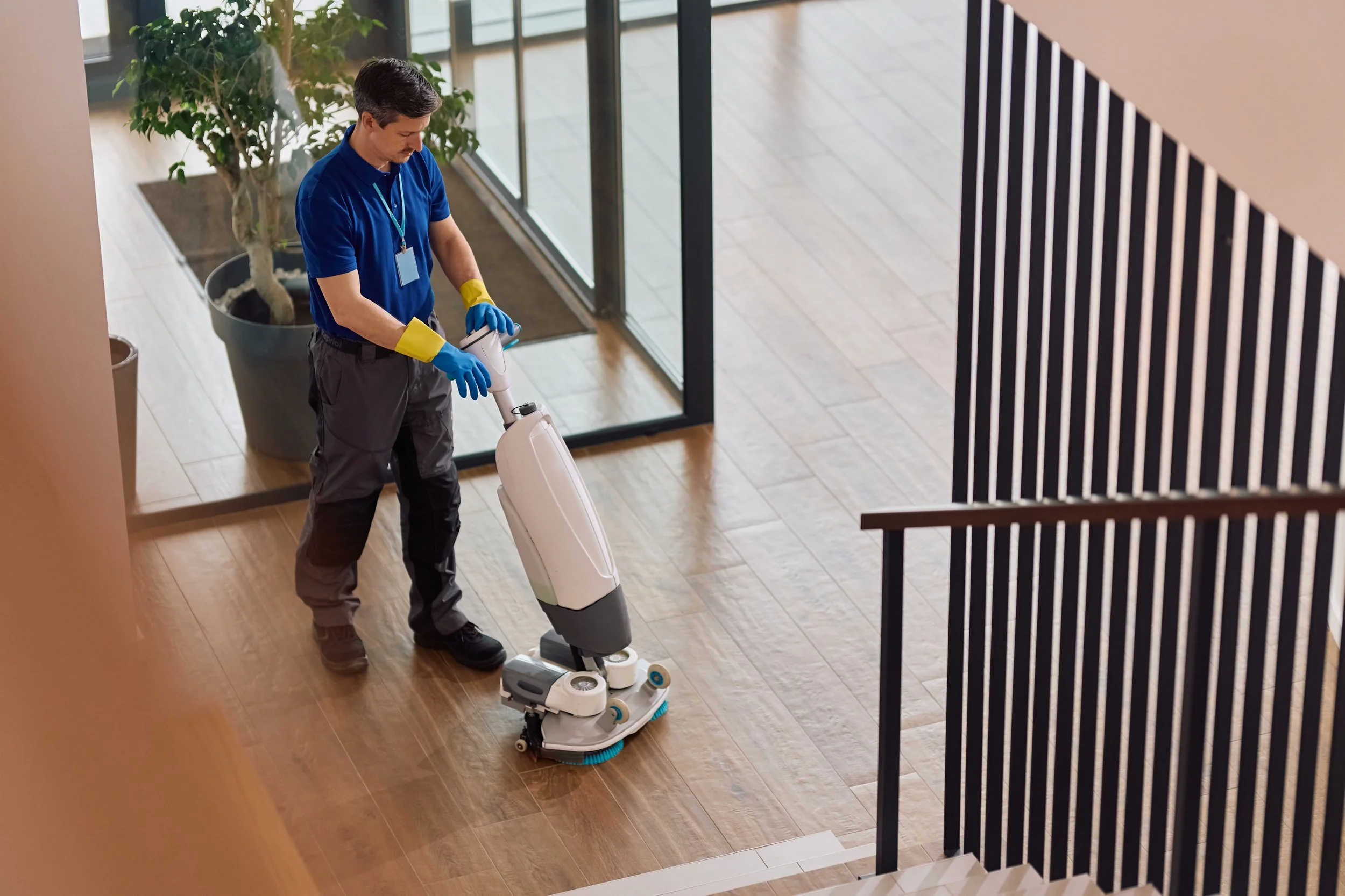 A man in a blue shirt and gray pants using a robotic floor cleaner in a modern building lobby with wooden flooring, large glass doors, and potted plants.