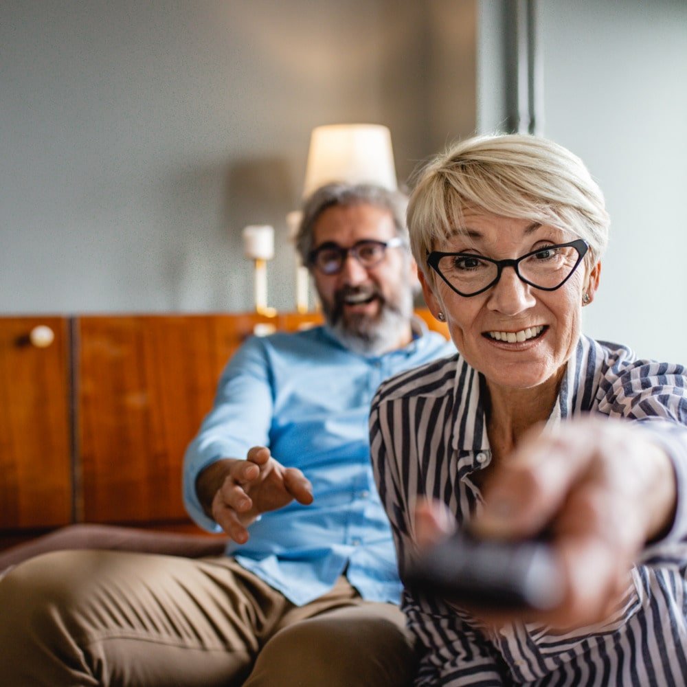 Une femme âgée avec des lunettes, souriante, tient une télécommande. Un homme âgé derrière elle, également souriant, semble lui parler ou faire un geste amical dans un salon lumineux.
