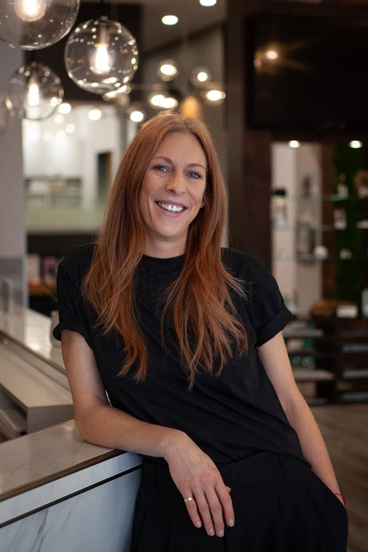 Une femme aux cheveux roux souriante, portant un t-shirt noir, posant dans un intérieur moderne avec des lampes en verre suspendues au plafond. Elle est audioprothésiste, service couvert ramq