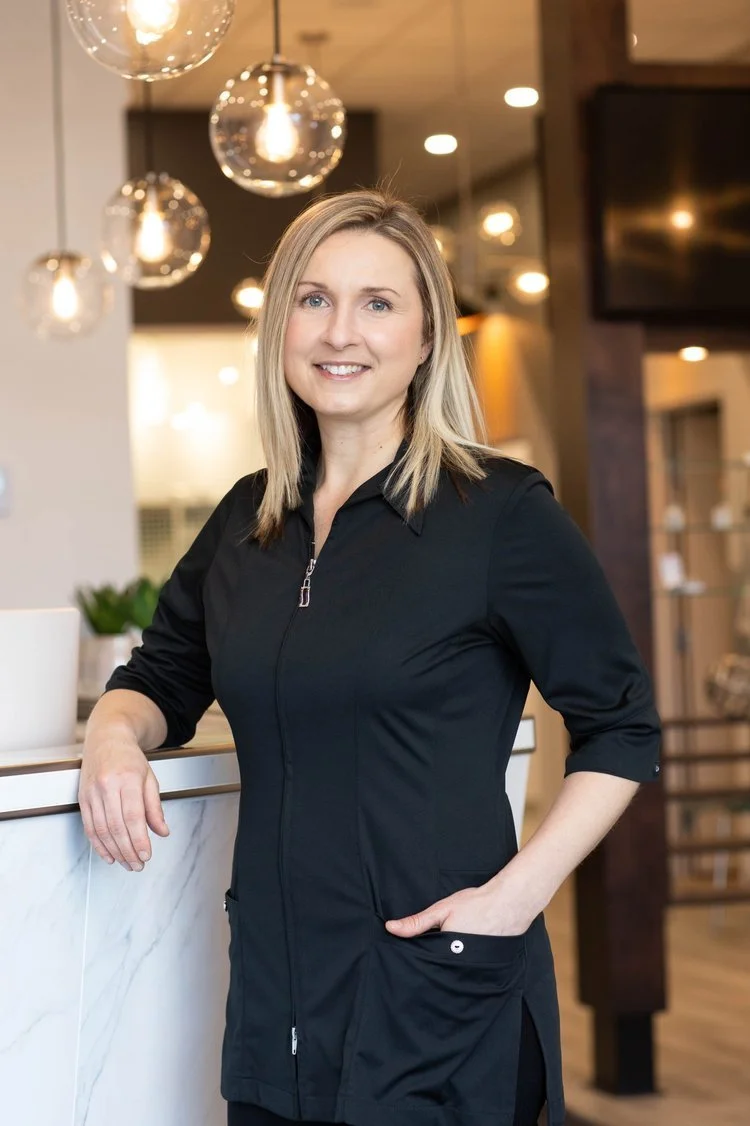 Femme blonde souriante, audioprothésiste, portant une veste noire dans un intérieur moderne avec des lampes suspendues