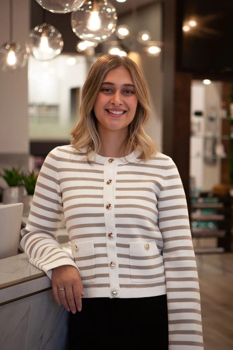 Jeune femme souriante avec cheveux blonds ondulés, portant un pull rayé beige et blanc avec des boutons décoratifs, dans un intérieur moderne avec éclairage suspendu