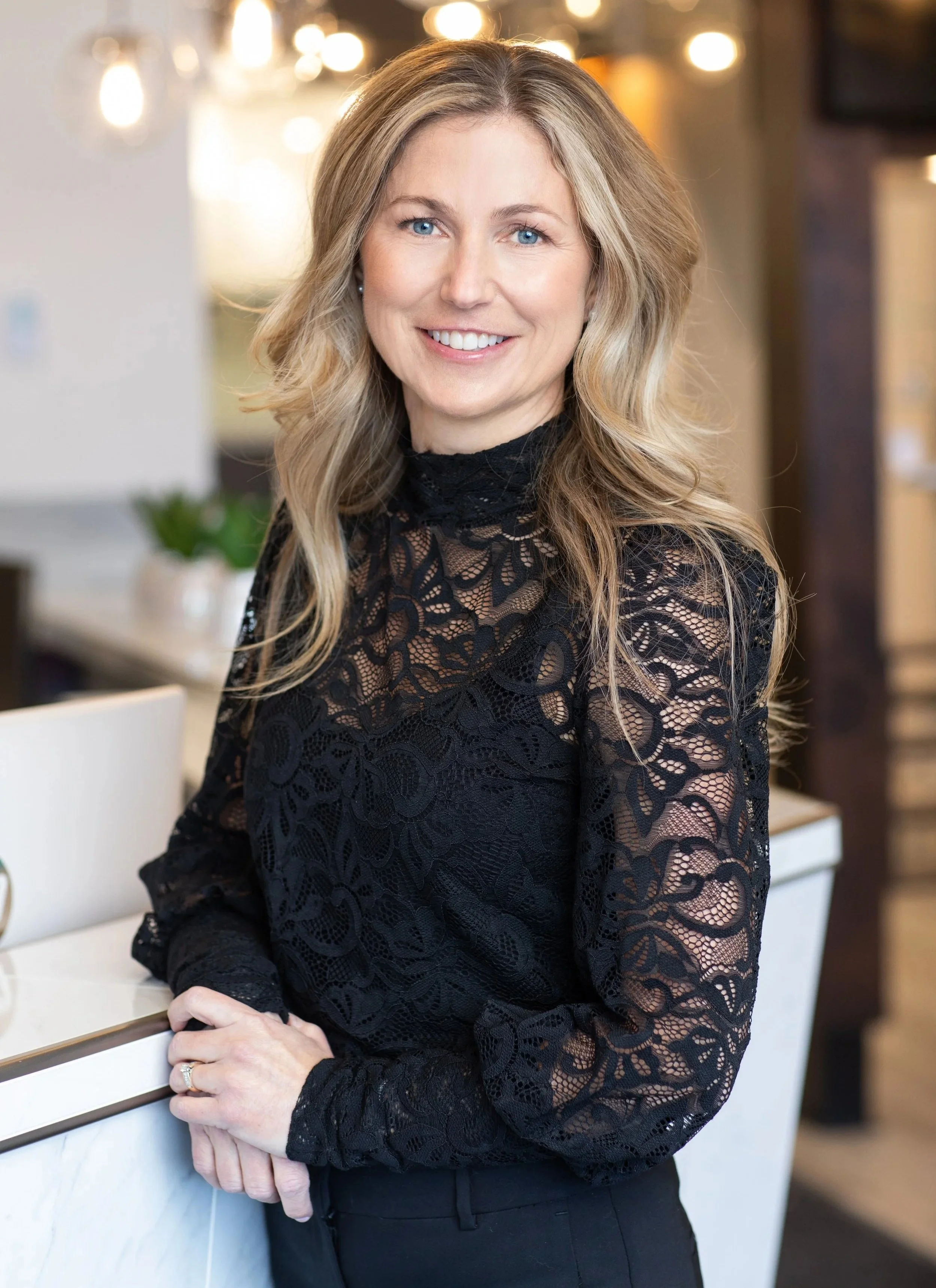Une femme souriante avec des cheveux longs et blonds, portant un haut en dentelle noire, posant dans un intérieur avec des lumières chaudes en arrière-plan. C'est la cofondatrice de la clinique Milot et Tremblay