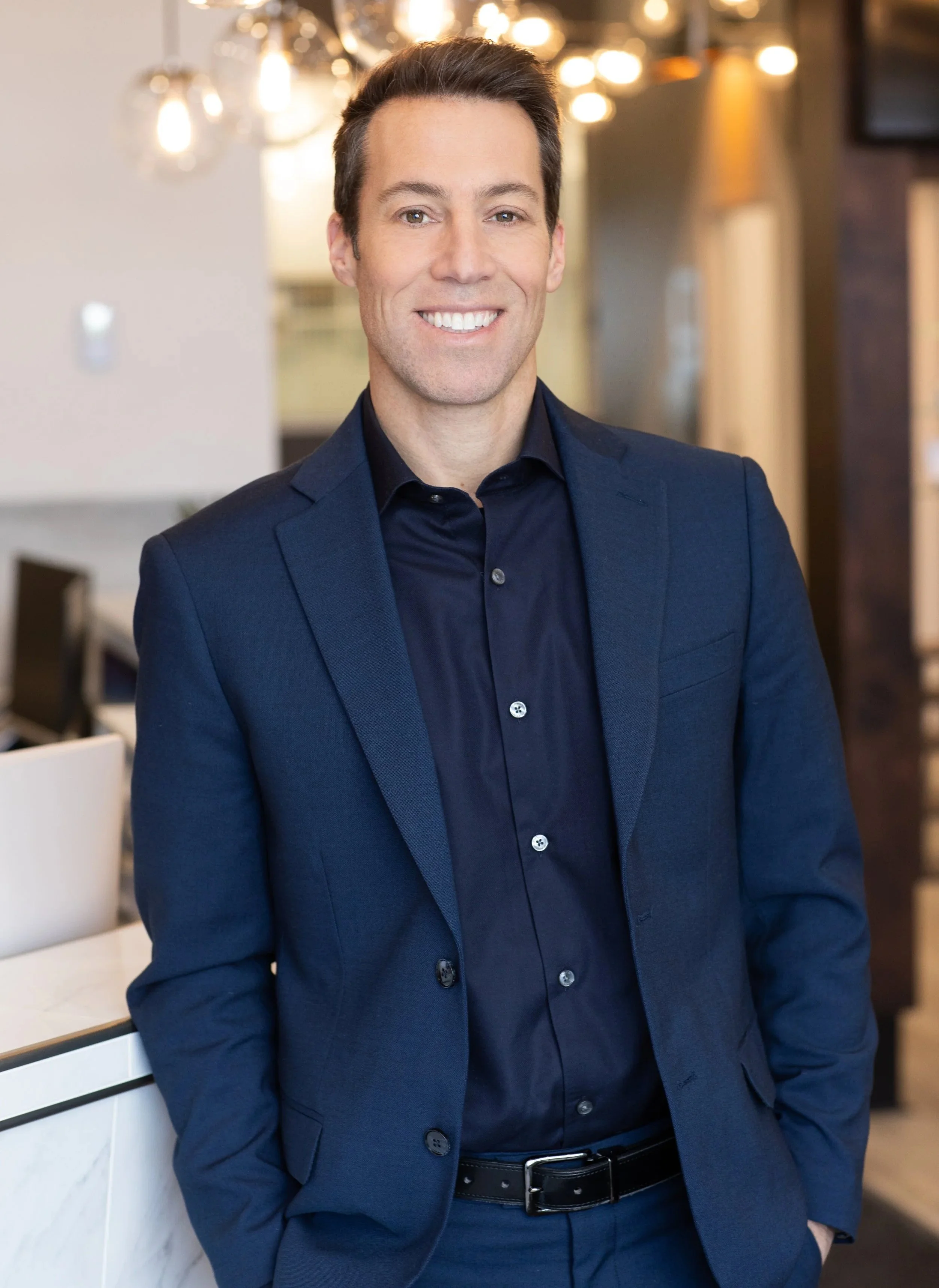 Un homme en costume bleu, souriant, dans un lieu intérieur moderne et bien éclairé. C'est le cofondateur de la clinique d'audioprothésiste