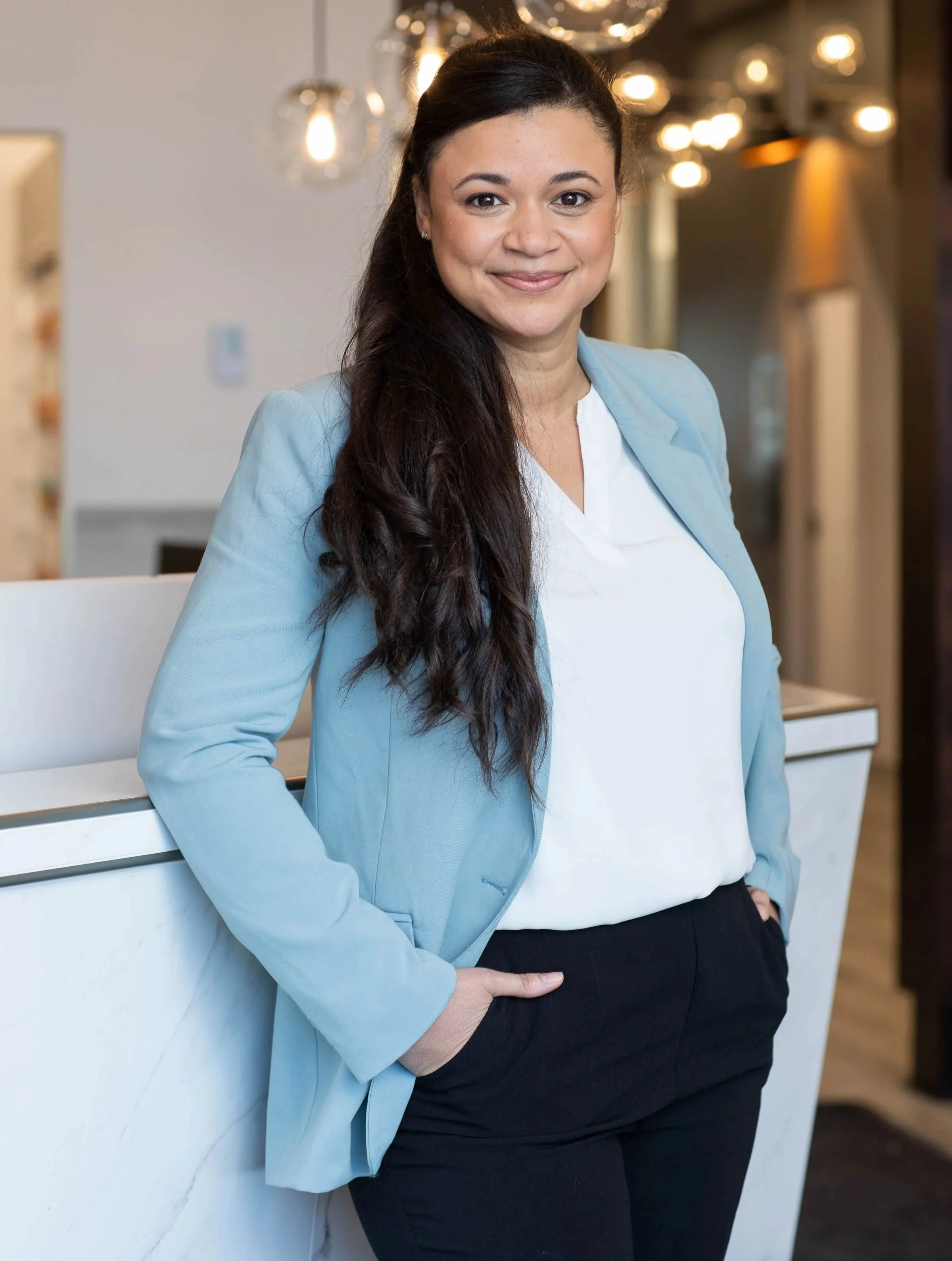 Une femme souriante portant une veste bleue et un pantalon noir, debout contre un mur blanc dans un environnement intérieur moderne avec des lumières suspendues. Elle est audioprothésiste