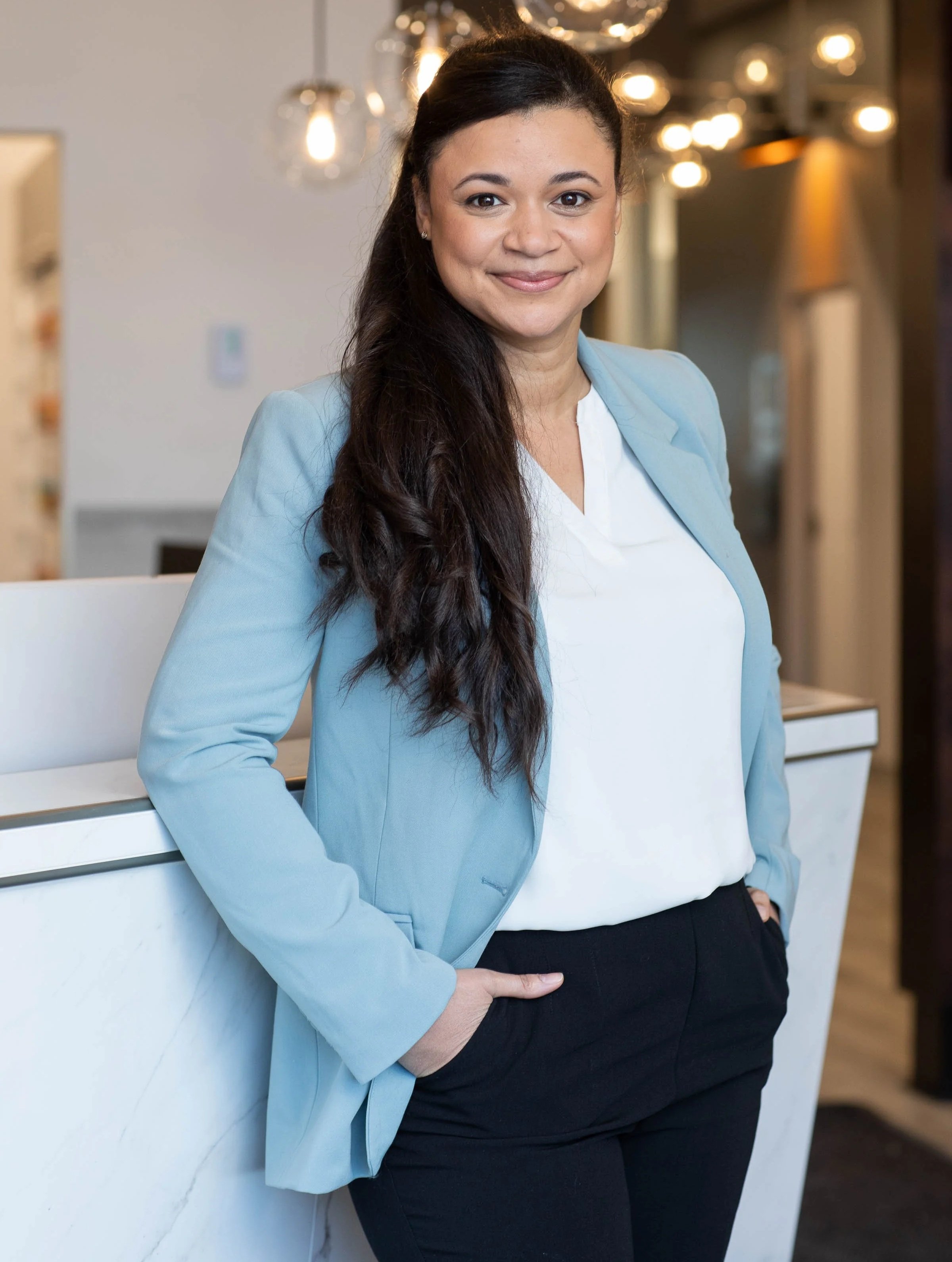 Une femme souriante portant une veste bleue et une chemise blanche, debout près d’un mur blanc, dans un intérieur moderne avec des lumières suspendues et un décor chaleureux. Elle est audioprothésiste et aide les personnes malentendantes.
