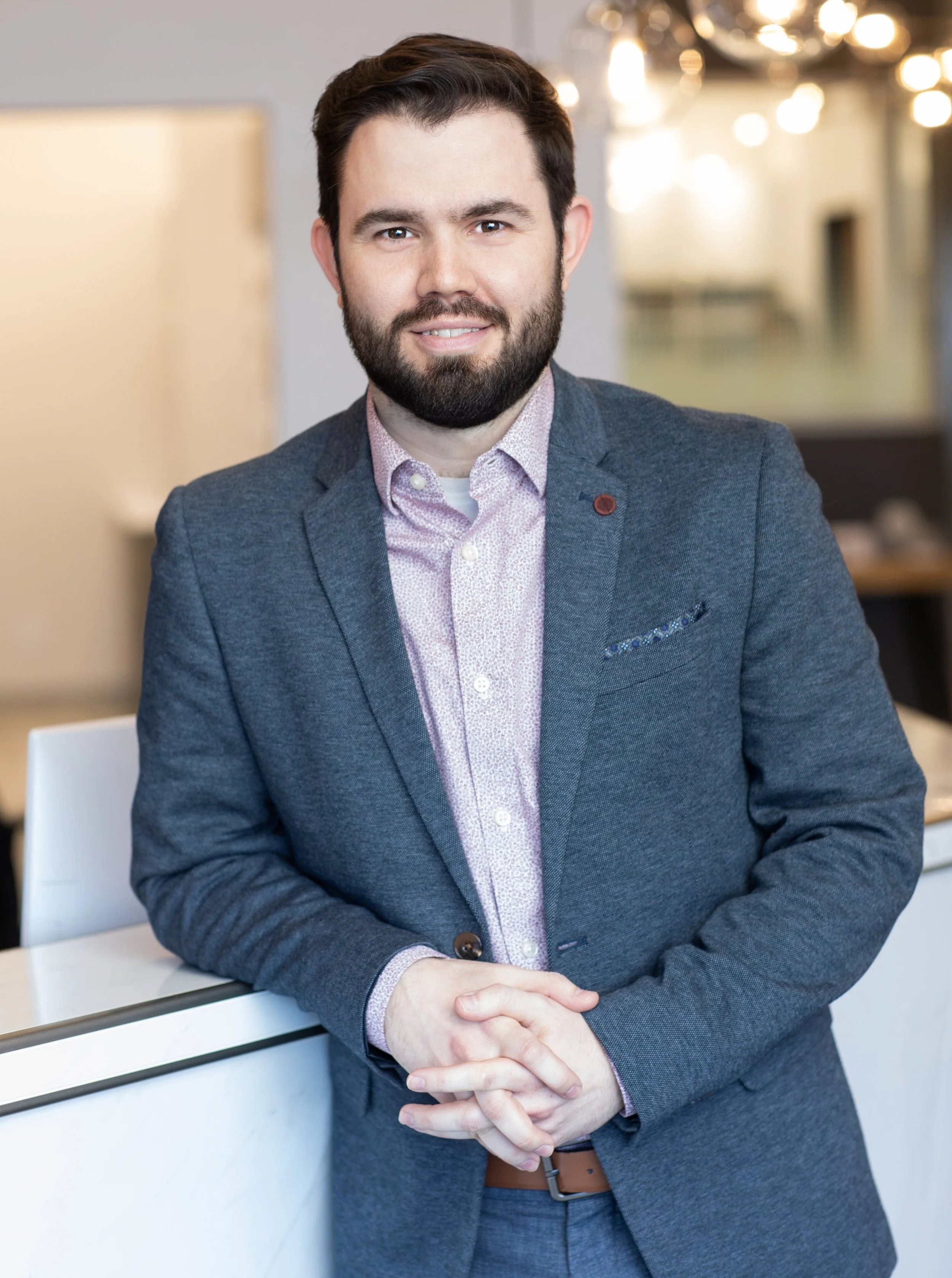 Portrait d'un homme d'affaires souriant, portant un costume bleu et une chemise rose clair, assis dans un bureau moderne.
