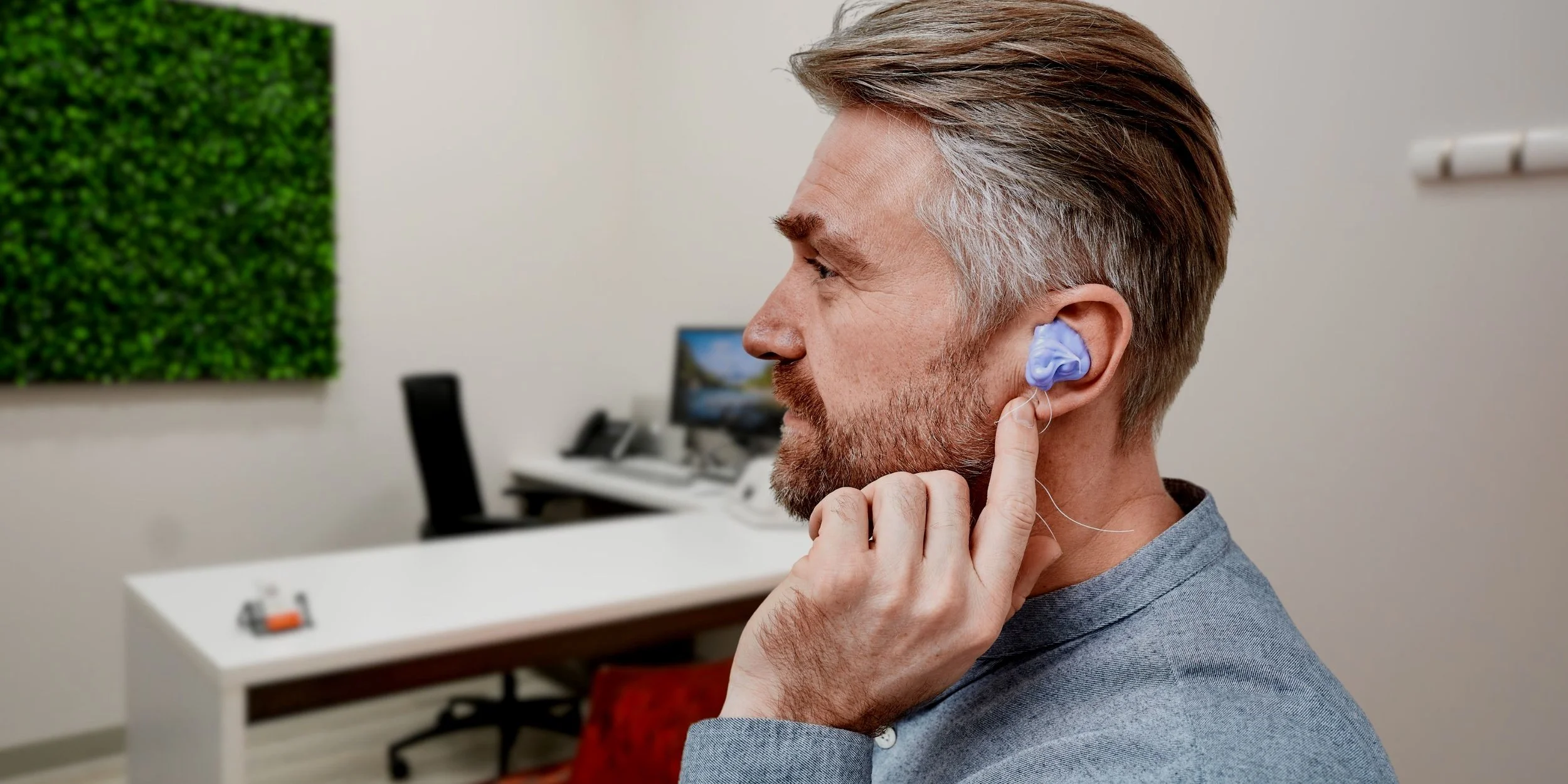 Un homme manœuvre une protection auditive avec son doigt dans un environnement de bureau, avec un fond comprenant un ordinateur, un téléphone, une peinture verte et un bureau blanc.