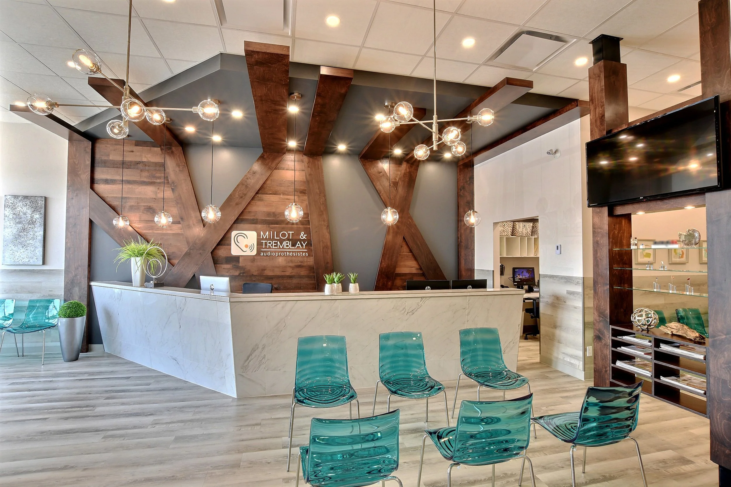 Intérieur d'une réception ou d'un salon avec un comptoir blanc, des chaises en plastique transparent turquoise, des lampes suspendues modernes, un mur en bois décoratif, une grande télévision à droite, et des plantes vertes.