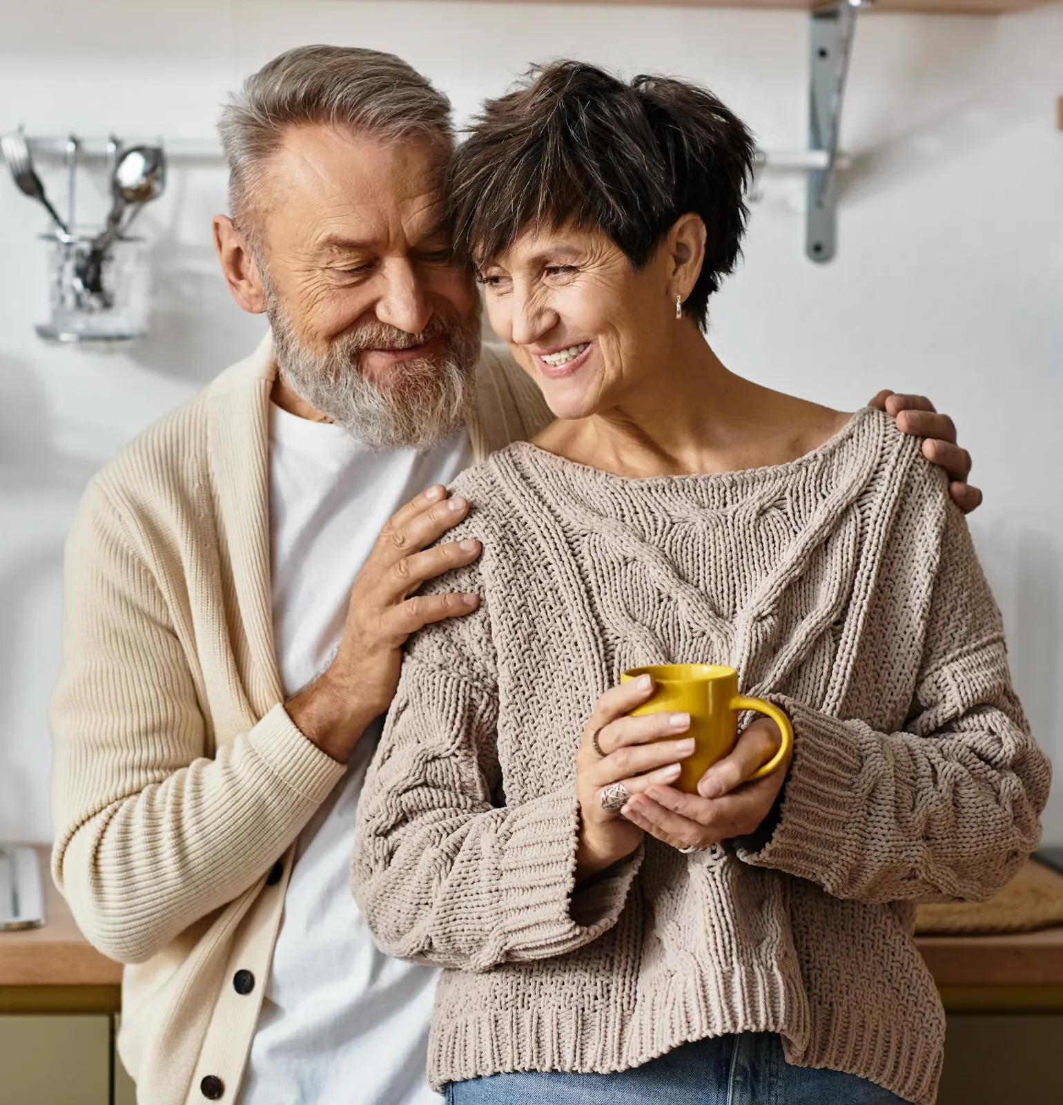 Un homme et une femme âgés partageant un moment de tendresse dans une cuisine, la femme tient une tasse jaune.