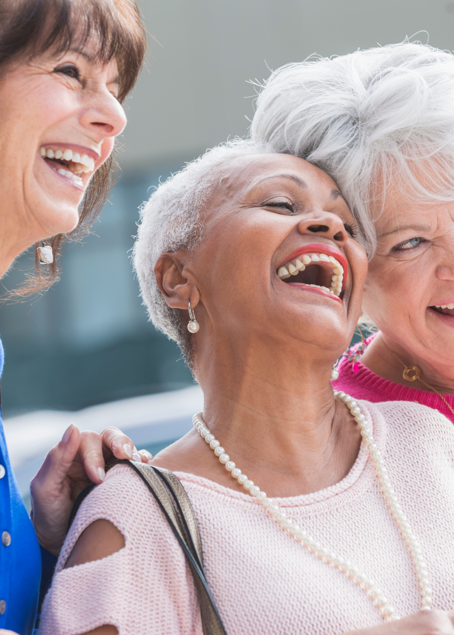 Trois femmes âgées rient et sourient ensemble, se tenant par l'épaule, dans un environnement lumineux et convivial. Elles ont bénéficié d'un crédit d'impot sur leurs appareils auditifs