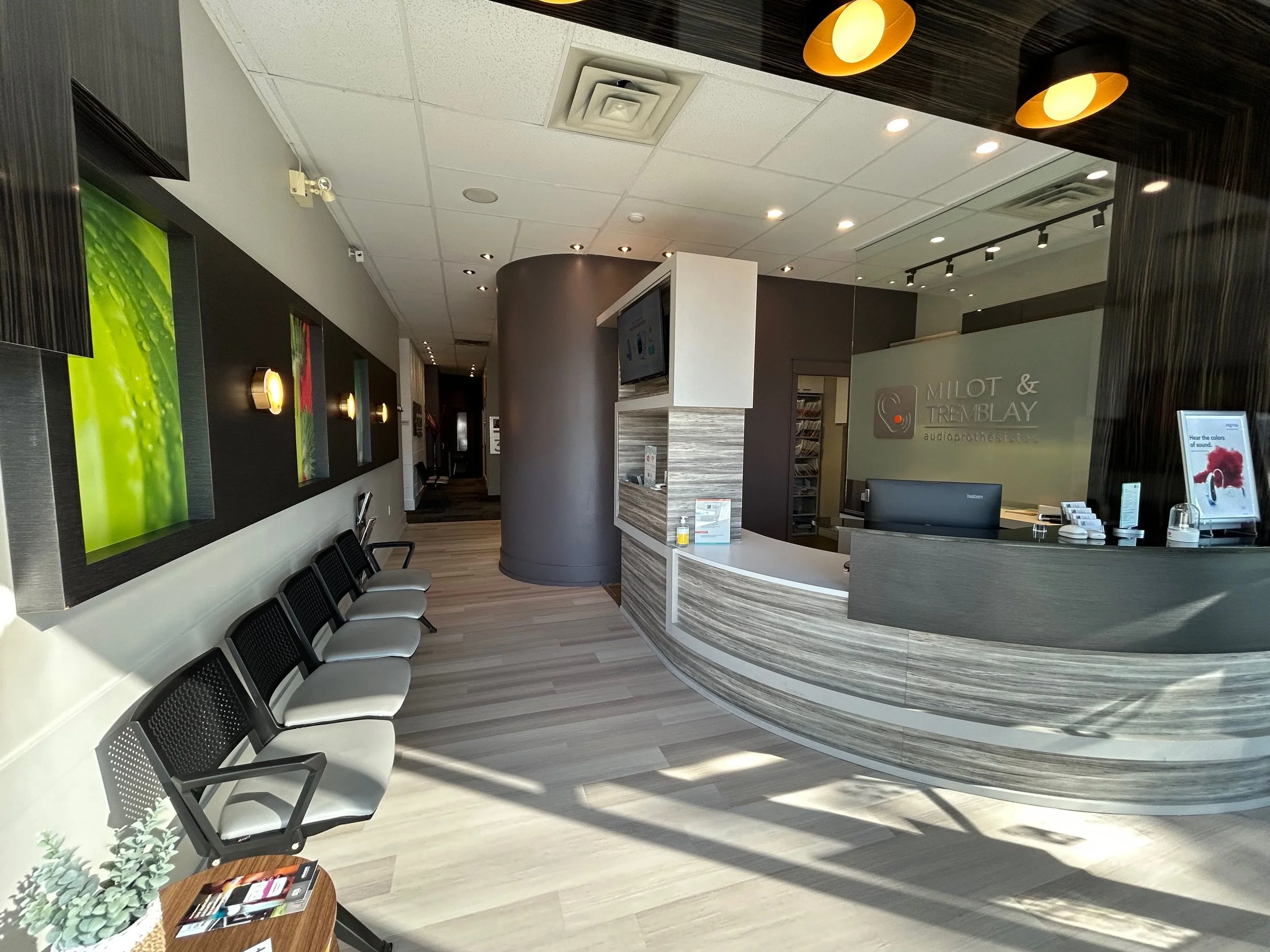 Salle d'attente d'une clinique d'audioprothésiste avec des chaises alignées contre le mur, décorée avec des œuvres d'art colorées, et un comptoir d'accueil avec un ordinateur et des brochures.