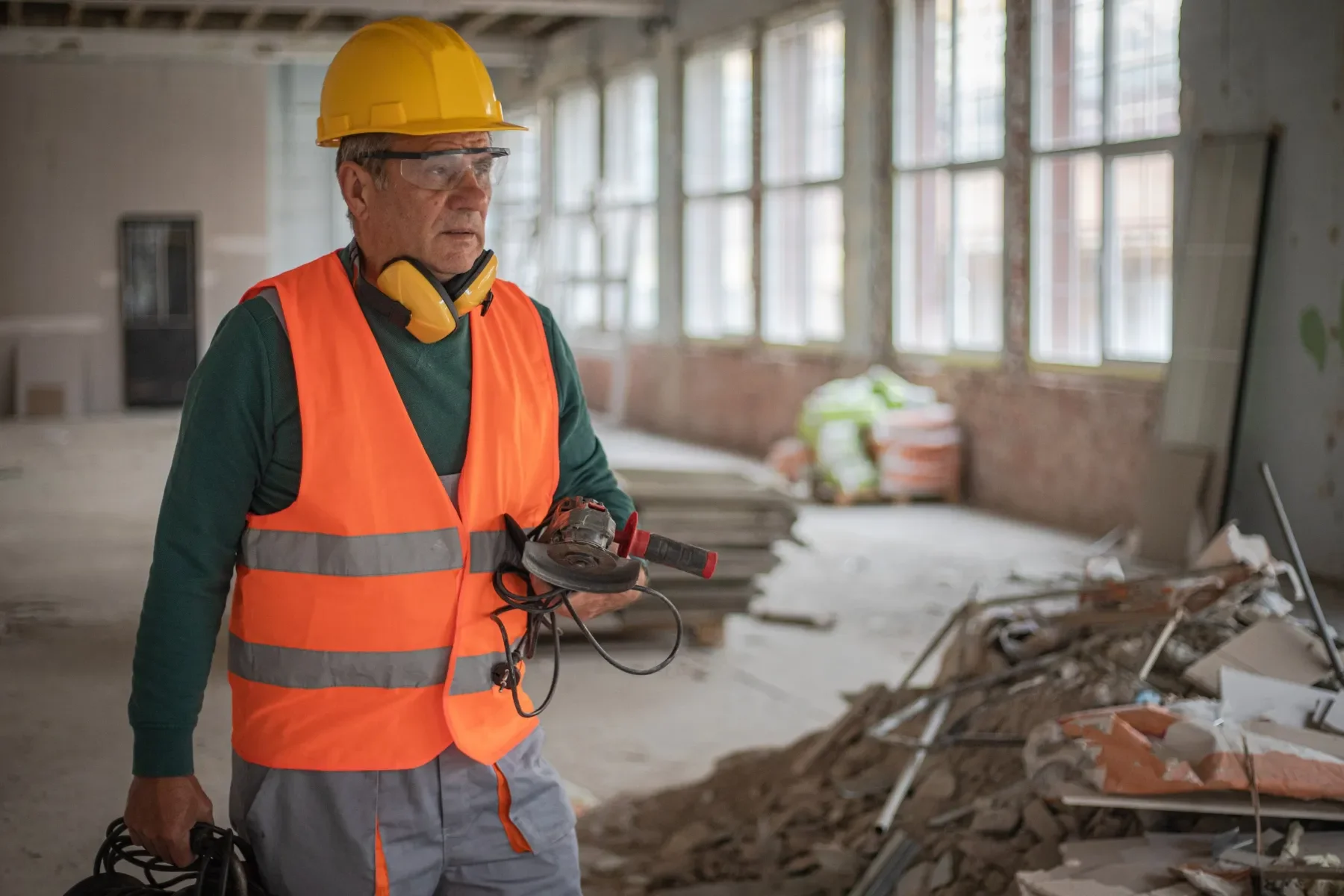 Un homme portant un casque de sécurité jaune, des protections auditives autour du cou, un gilet de sécurité orange et gris, regarde un écran dans un bâtiment en cours de rénovation, avec des débris et des matériaux de construction autour.