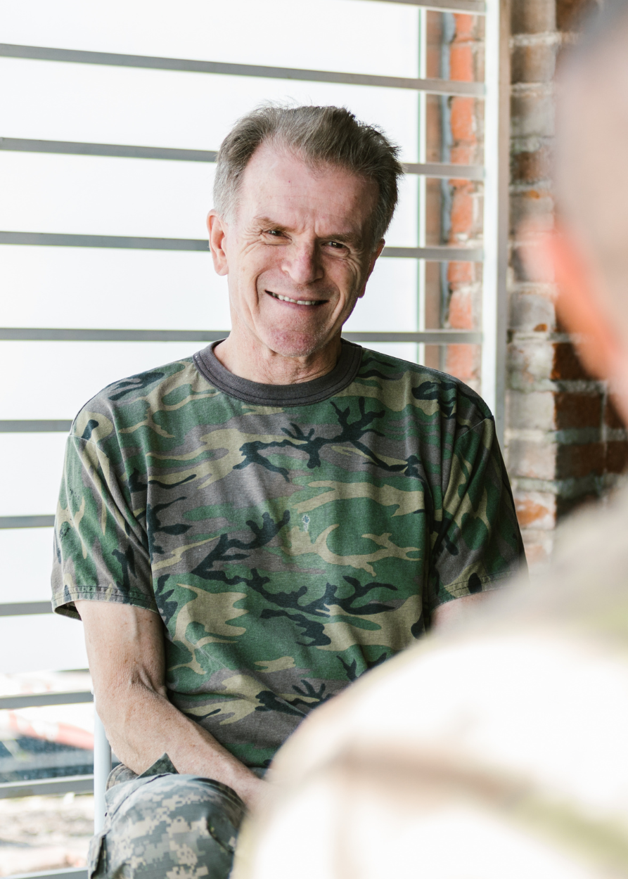 Un homme, ancien soldat souriant avec des cheveux gris, portant un t-shirt camouflage vert et une tenue camouflage, assis à l'extérieur près d'un mur en briques avec des volets blancs en arrière-plan.