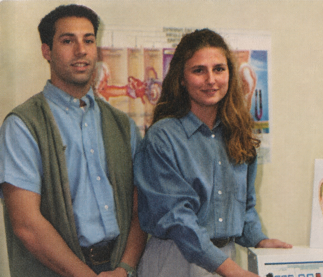 Deux jeunes adultes, un homme et une femme, posent devant un tableau biologique dans une salle de classe.
