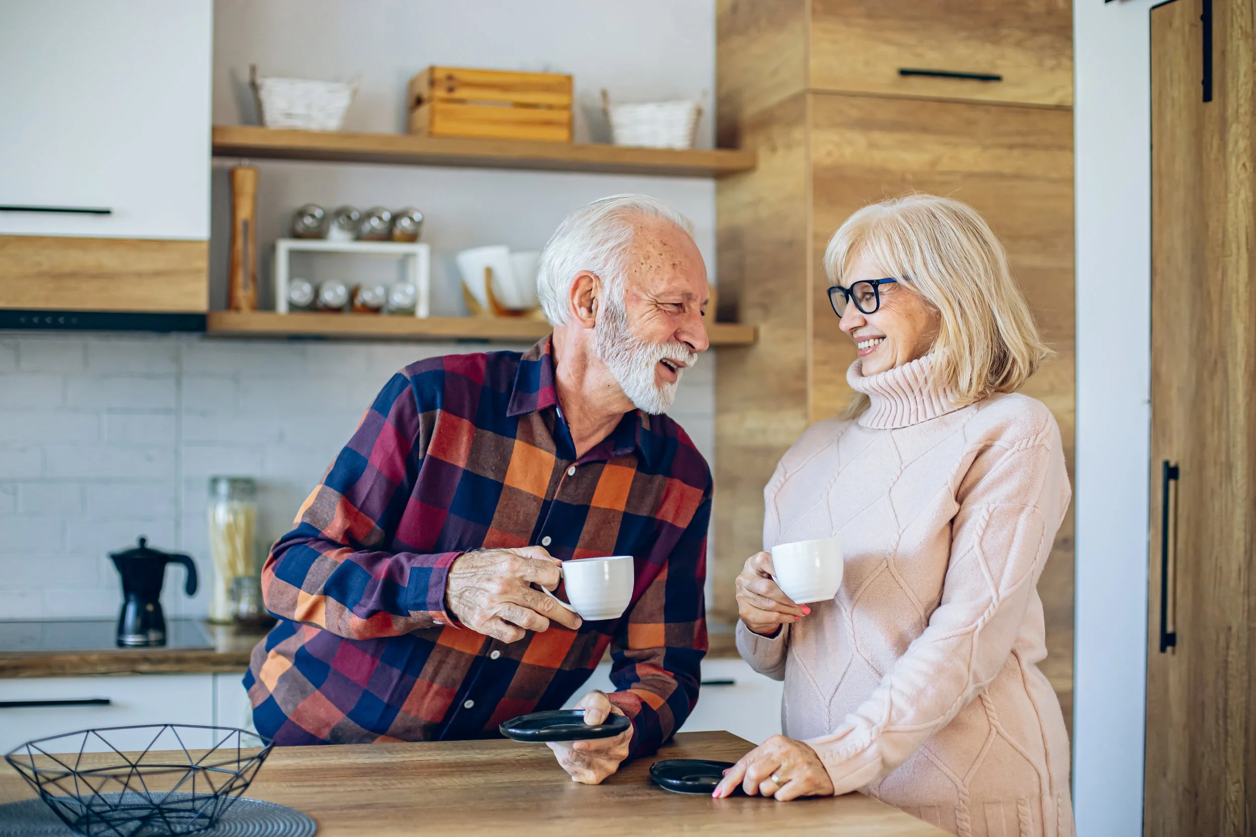 Un homme âgé et une femme âgée sourient en partageant une tasse de café dans une cuisine moderne en bois.