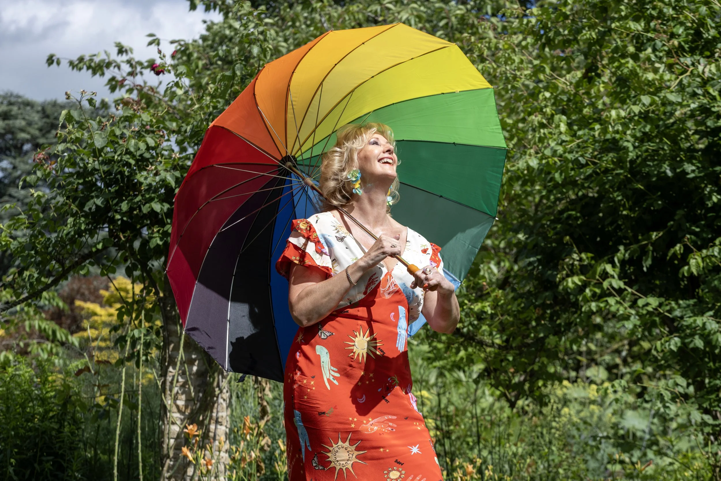 A woman holding a colorful rainbow umbrella outdoors surrounded by greenery, smiling and looking up.