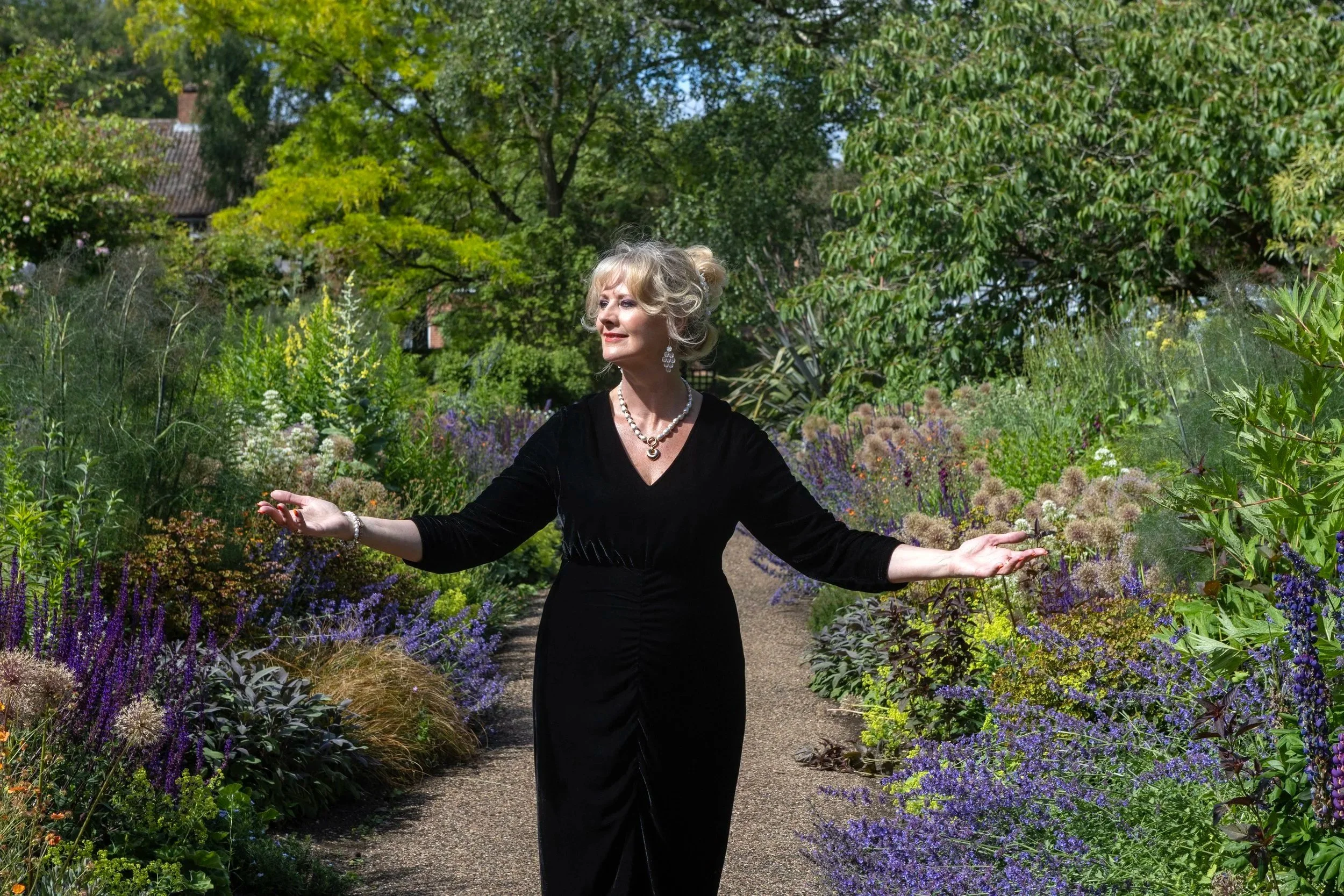 A woman in a black dress standing with her arms outstretched in a lush garden filled with colorful flowers and green trees.