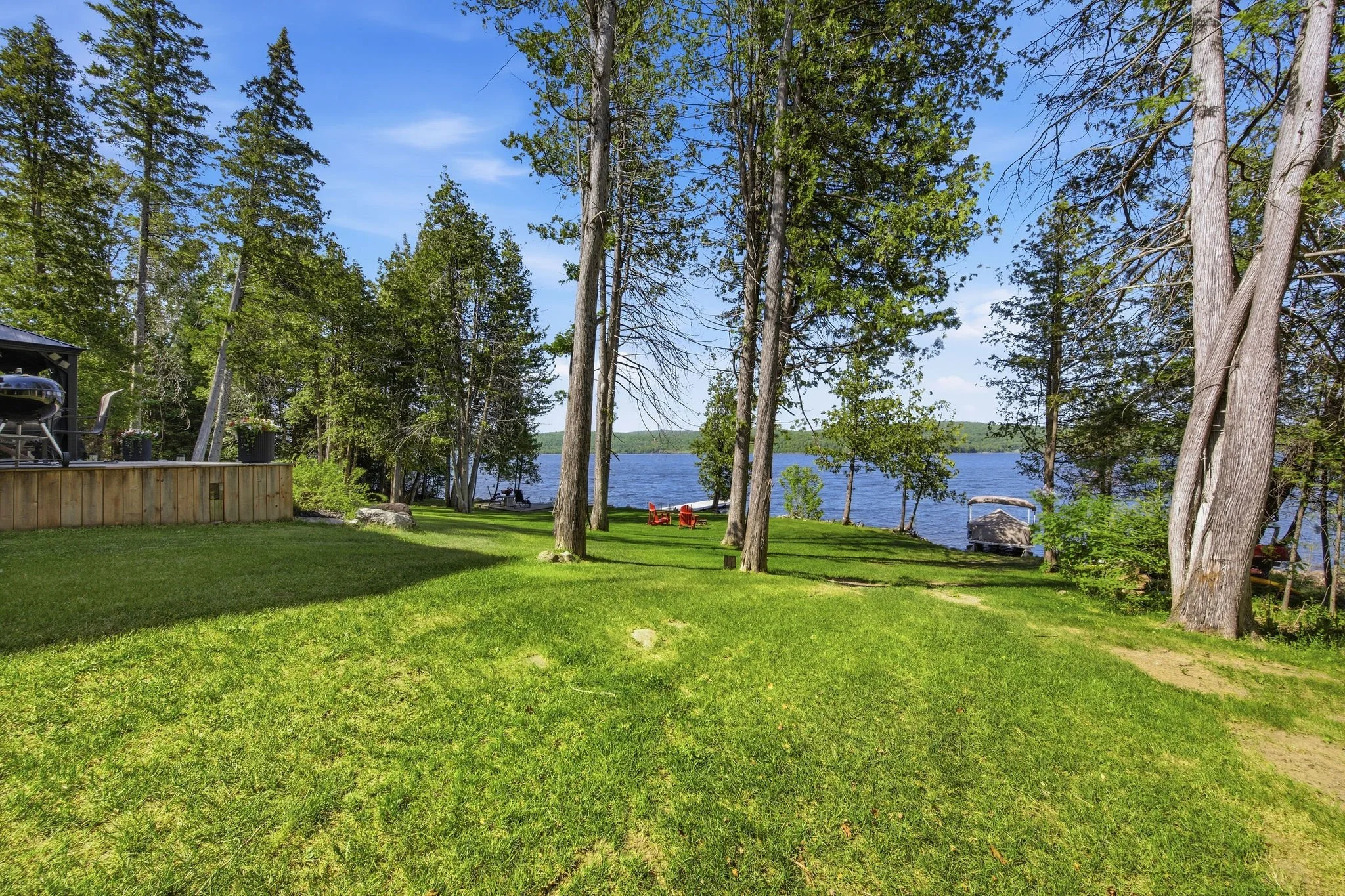 Lush green backyard with tall trees, a wooden deck, a body of water in the background, and red Adirondack chairs.