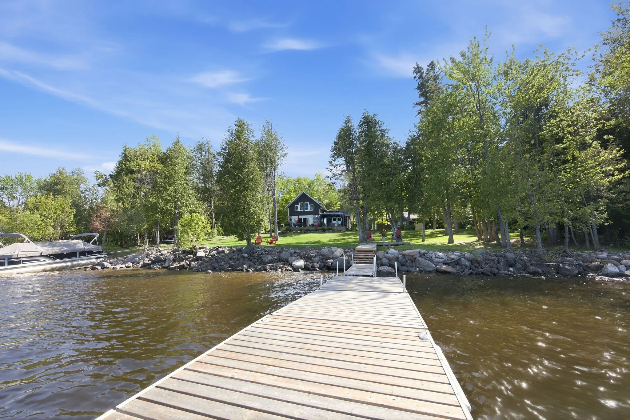 A wooden dock extends over a lake towards a grassy yard with trees and house, under a blue sky with wispy clouds.