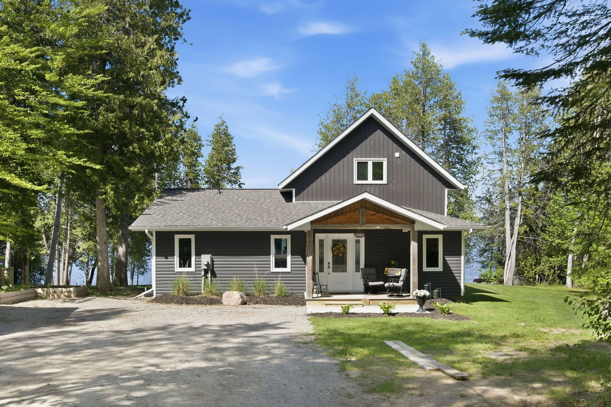 A modern, two-story gray house with white trim and a front porch, surrounded by green trees under a blue sky.