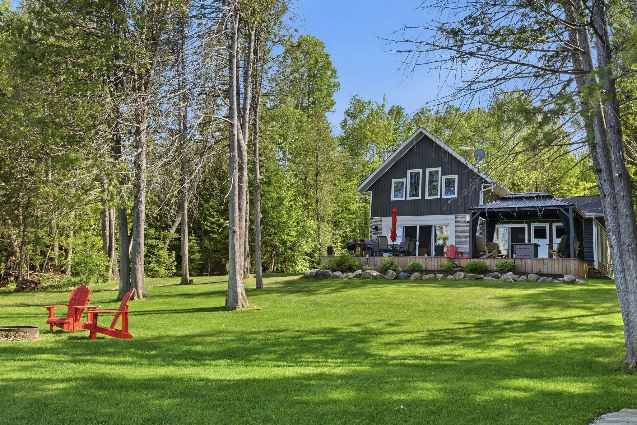 A backyard with green grass, red Adirondack chairs around a fire pit, tall trees, and a modern black house with large windows and an outdoor deck.