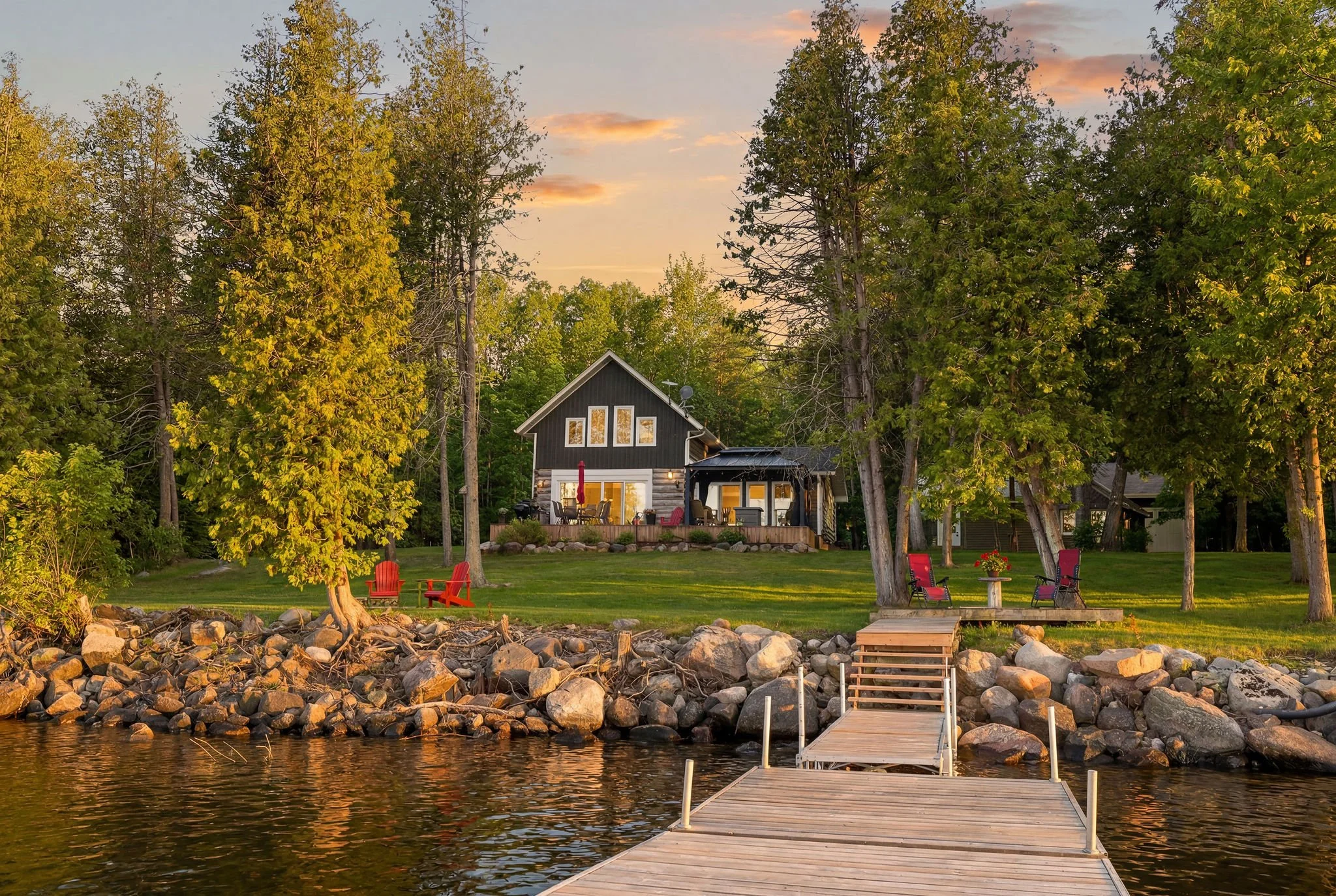 A lakeside backyard scene at sunset with a wooden dock extending into the water, surrounded by rocks and green grass. There are red Adirondack chairs and a picnic table on the lawn, and a large house with a deck and bright lights is nestled among tal