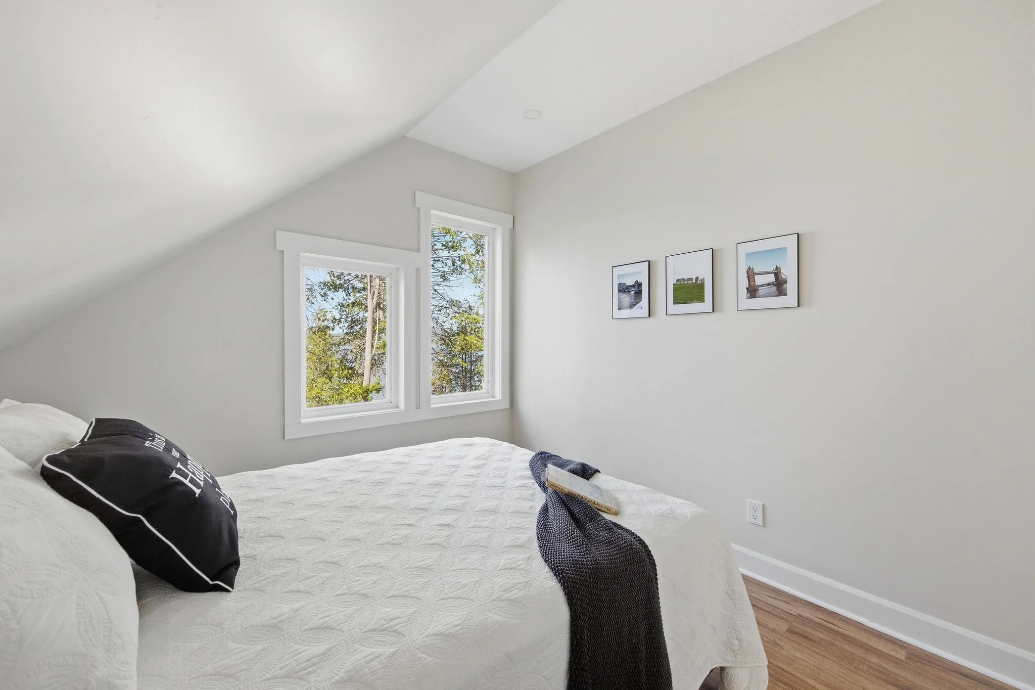 A bedroom with white walls, a white quilted bedspread, and black decorative pillows. There are two windows with white trim, showing trees outside. Three framed photographs of bridges are hung on the wall above the bed. A pair of gray slippers and a b