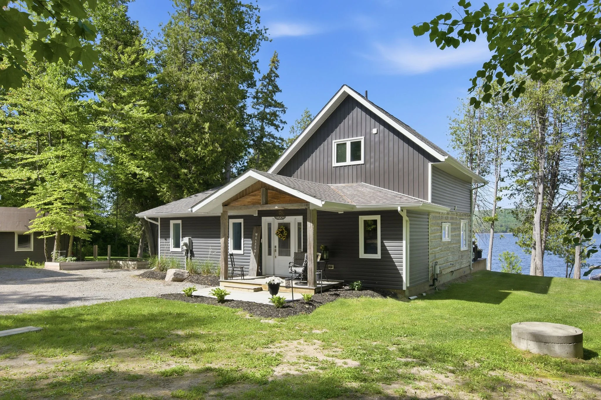 A modern two-story house with gray siding and white trim, surrounded by tall green trees, near a body of water, with a front porch and outdoor seating.
