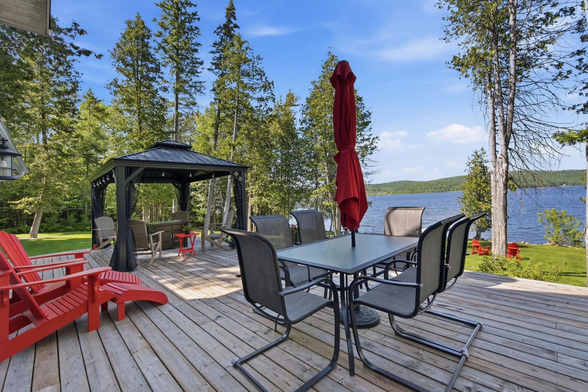 A spacious outdoor deck by a lake with trees, a gazebo, outdoor furniture including chairs, tables, and a red umbrella, and a scenic view of the water and distant hills.