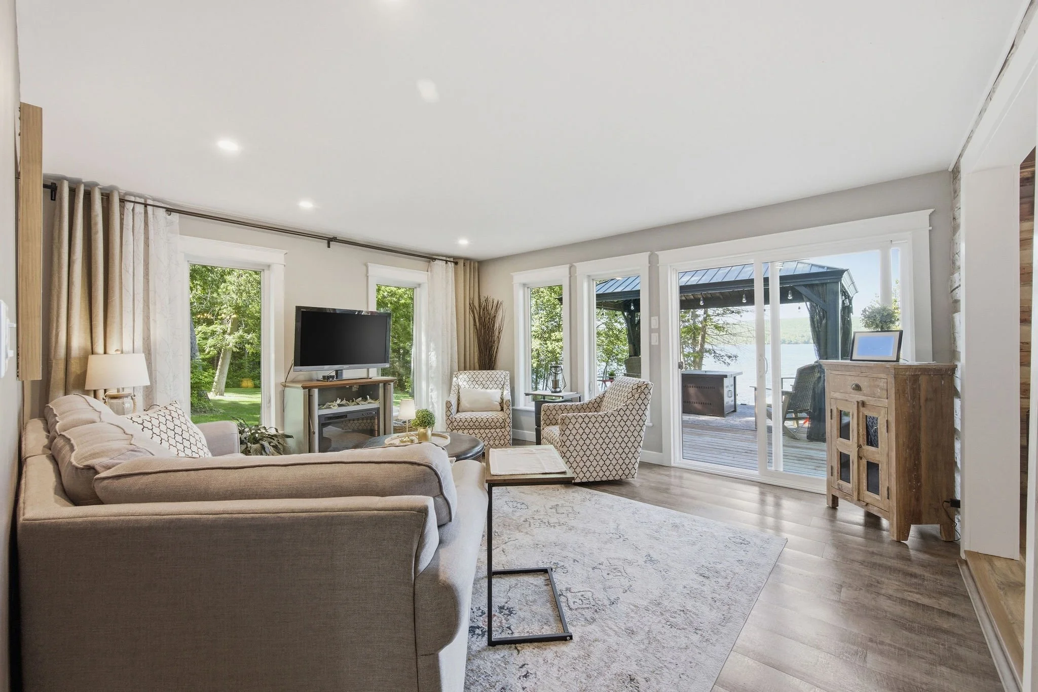 Living room with large windows showing a view of green trees and water, beige sofa, armchairs, coffee table, TV, and wooden furniture, with sliding glass doors leading to an outdoor deck.
