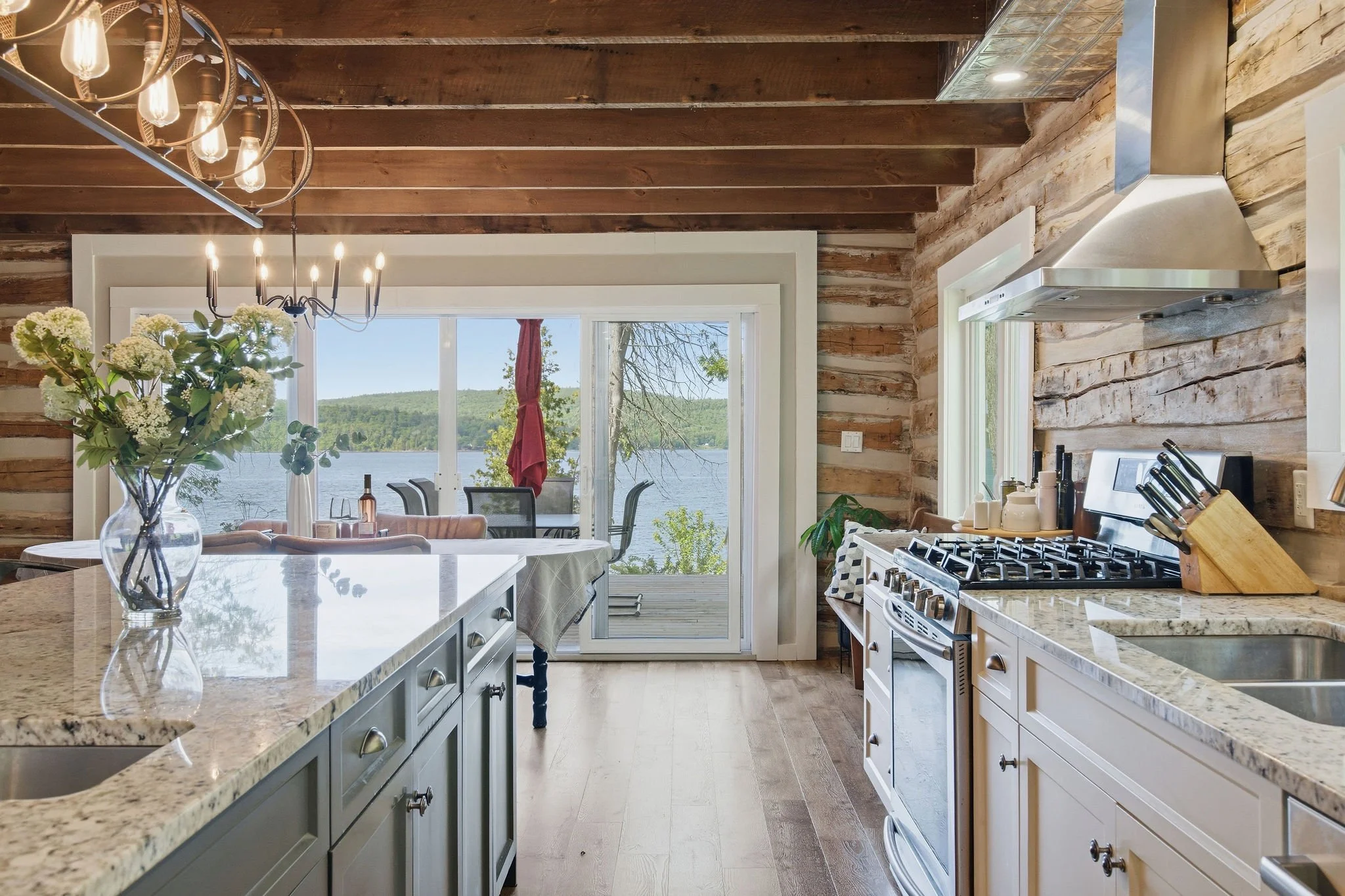 A view from the kitchen looking out onto a house's backyard with a lake, trees, and a mountain in the distance through large sliding glass doors. The kitchen has wood-paneled walls and a wood-beamed ceiling, with a chandelier hanging above the dining