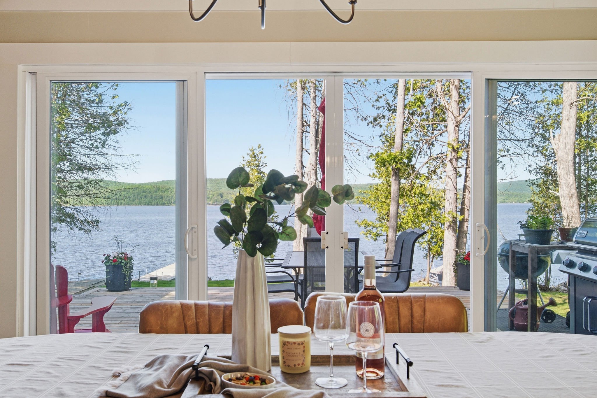 View through large sliding glass doors shows a lakeside outdoor patio with chairs and greenery, overlooking a body of water and trees under a clear blue sky.