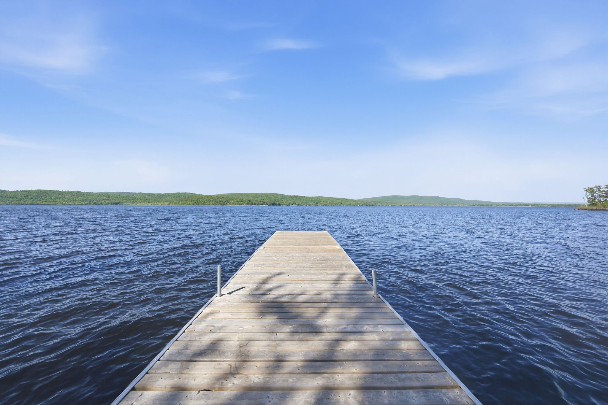 A wooden dock extends into a large body of water, with a view of distant green hills and a blue sky with few clouds.