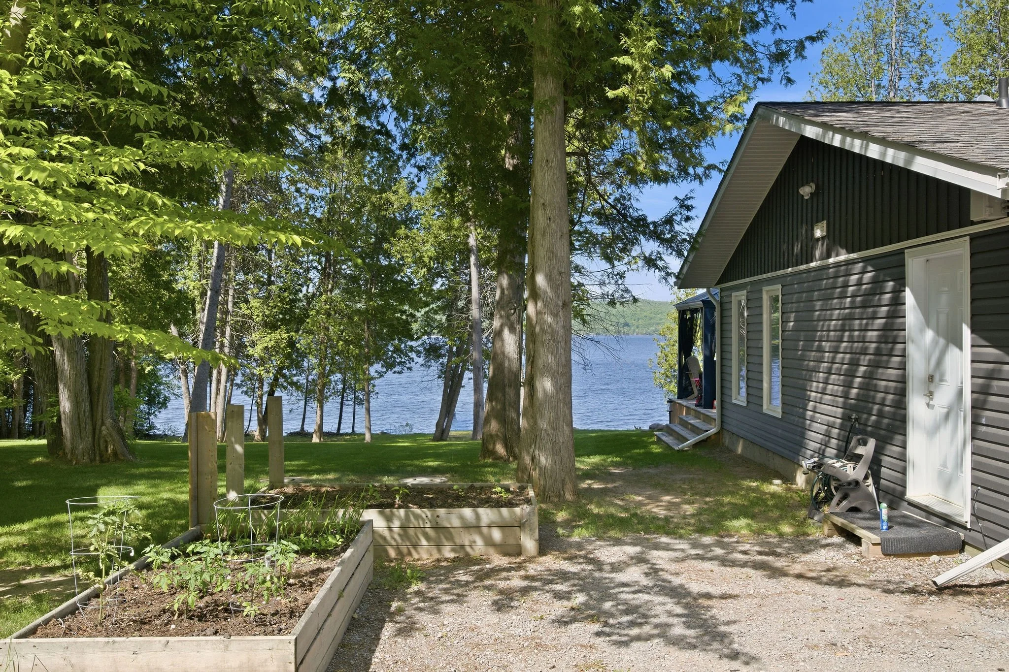 A backyard garden near a lake with green grass, trees, a raised garden bed, and a gray house with a slanted roof, windows, and steps leading to an open door.