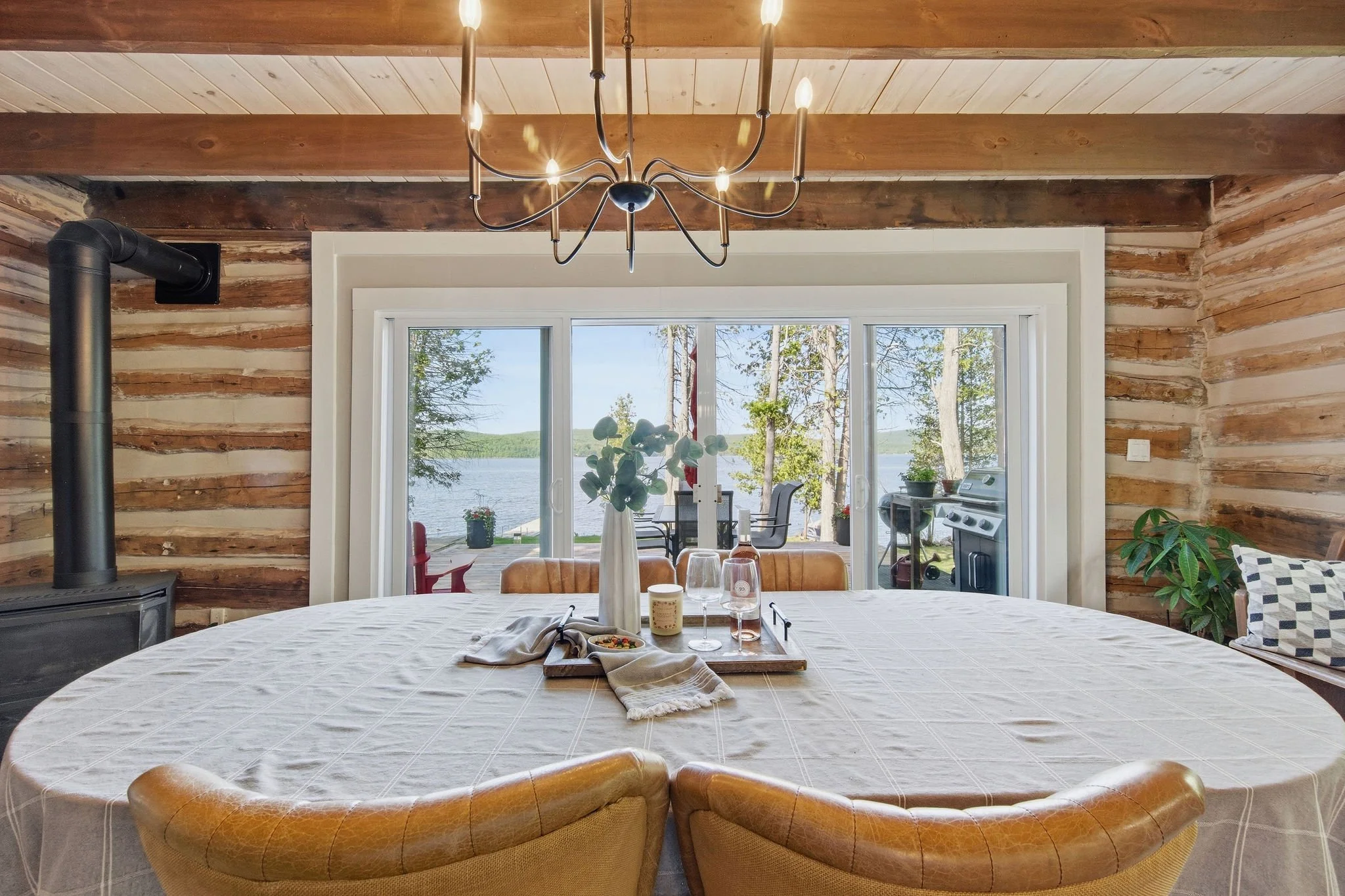 A dining room with a round table covered with a white tablecloth, set with drinks and a tray of food, and surrounded by tan leather chairs. The room features rustic wooden walls and a ceiling with exposed beams. A large window reveals a lakeside view