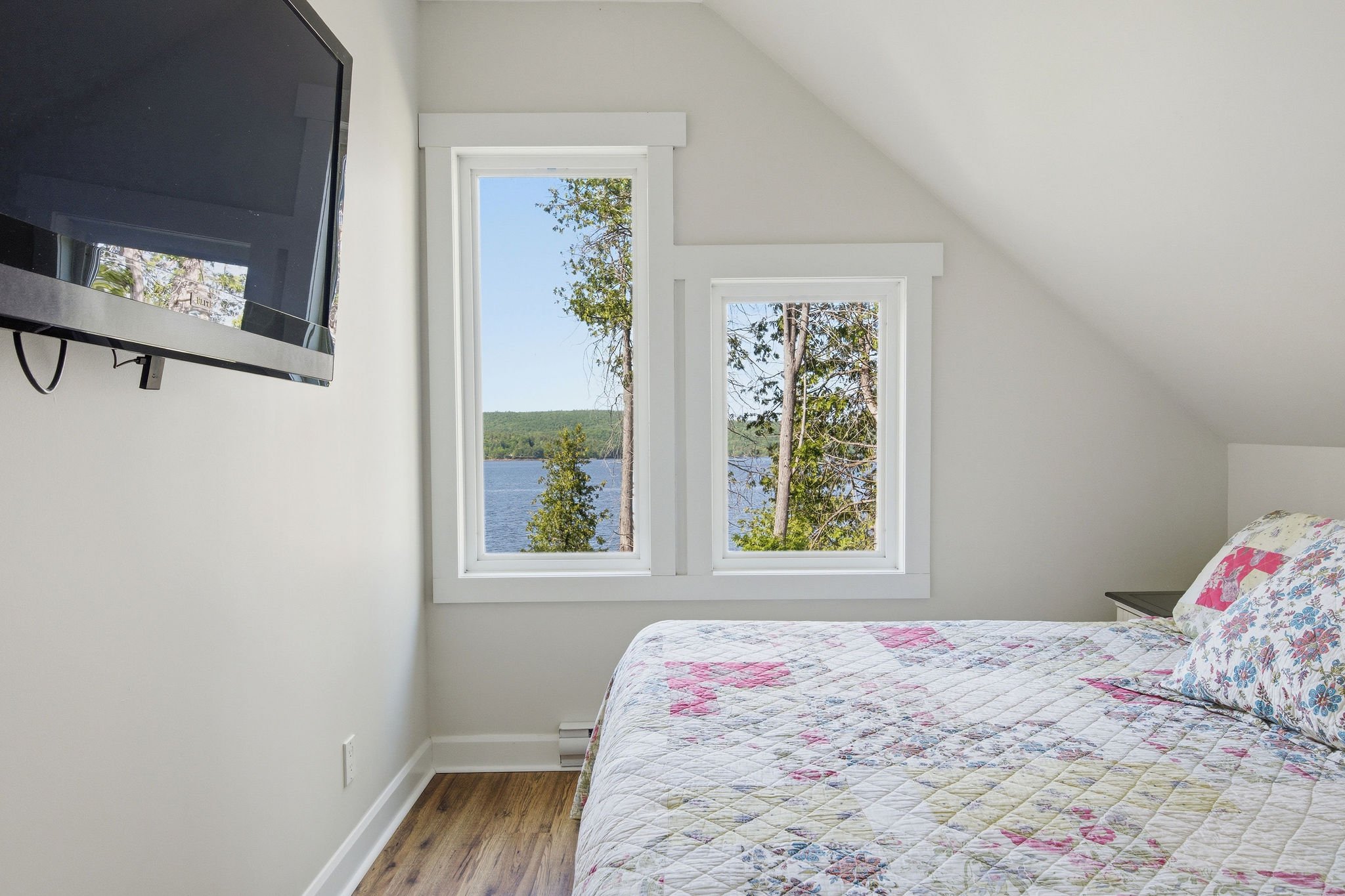 A bedroom with a white wall, a bed with a quilted floral blanket, and a window with a beautiful view of trees and a lake outside.