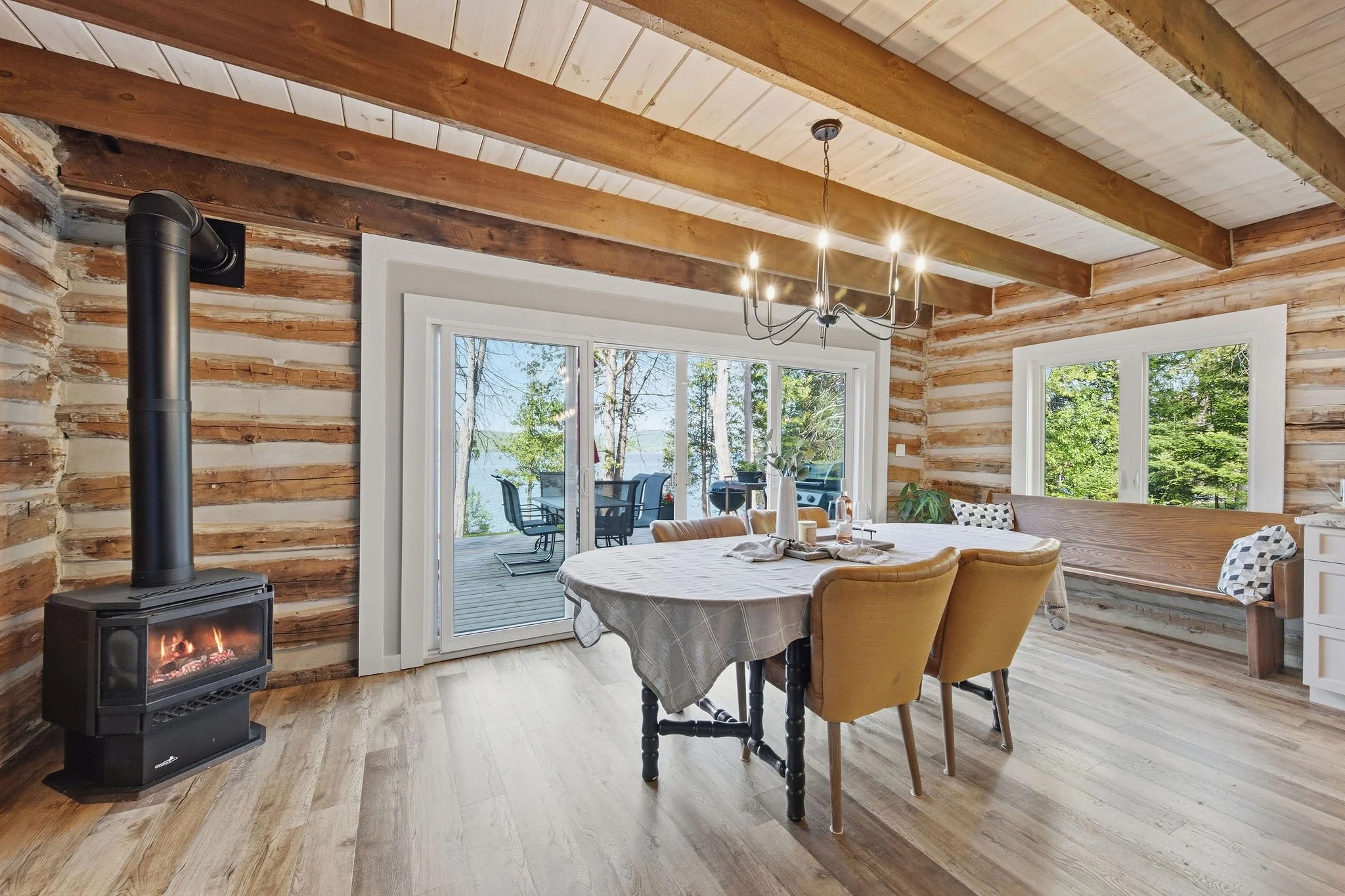 Interior of a rustic log cabin dining area with a round table, yellow chairs, a wood-burning stove, large windows, and a view of a deck with outdoor furniture and trees outside.