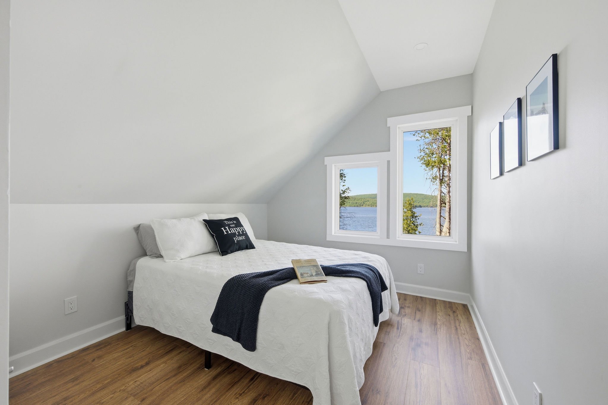 A bedroom with white walls, a bed with white bedding, a black pillow that says 'This is our happy place,' and a navy blanket. There are two windows showing trees and a lake outside, and three framed pictures on the wall.