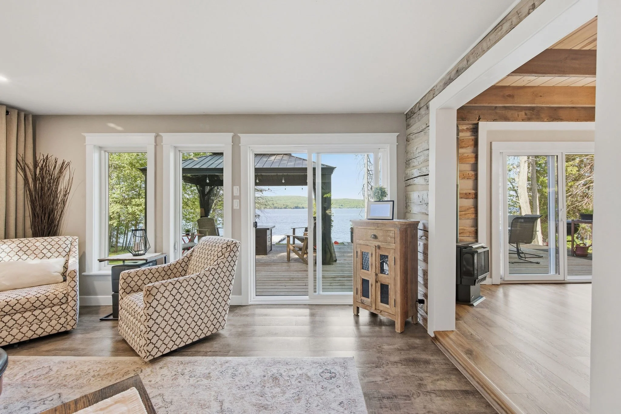 Living room with large windows overlooking a lake, with indoor furniture including patterned armchairs and a wooden cabinet, and a deck outside with outdoor furniture.