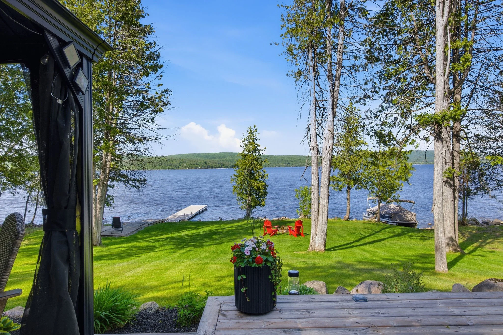 View of a lakeside scene taken from a porch with green grass, trees, a dock extending into the water, and a boat under a canopy. The sky is clear and blue.