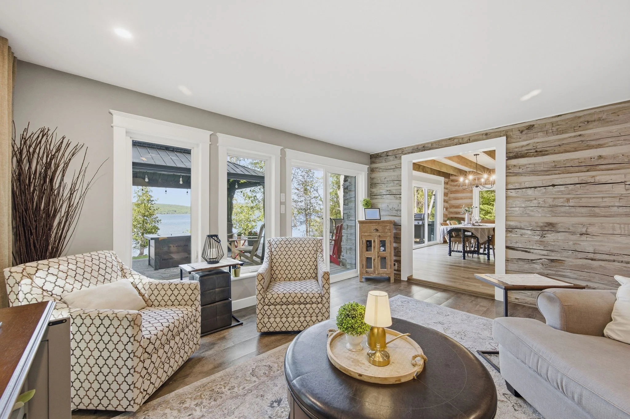 Living room with large windows, wooden accents, and furniture including patterned armchairs, a coffee table, and decor items.