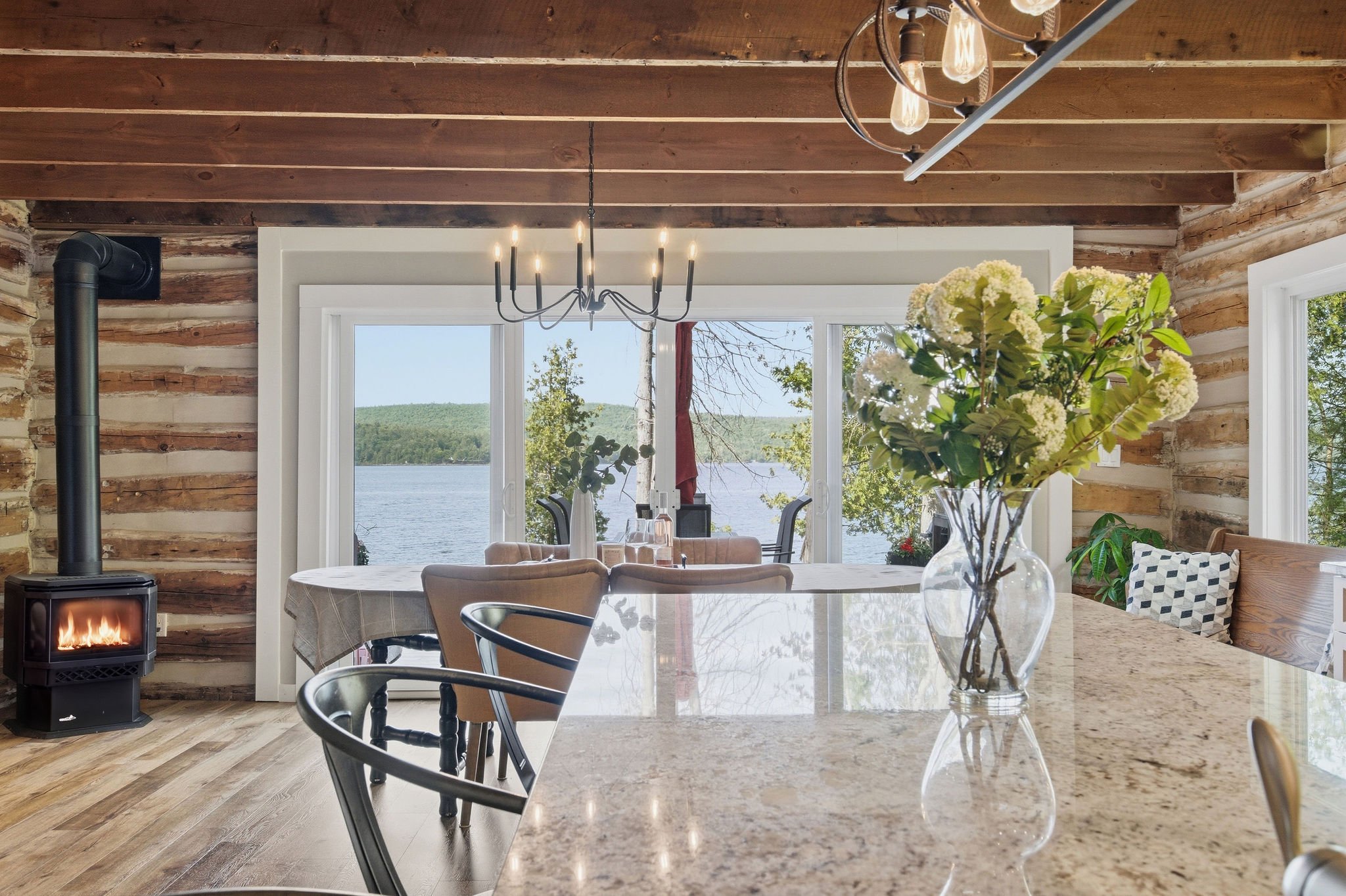 Interior of a rustic home with wood log walls, a large window overlooking a lake with trees, a chandelier, a vase of white flowers on the table, and a small fireplace
