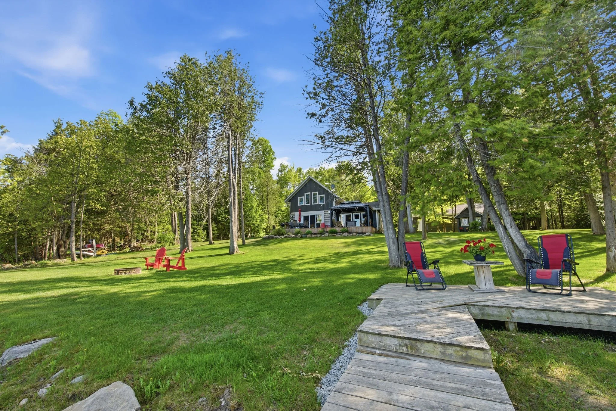 A backyard with green grass, tall trees, a wooden deck with chairs and a small table with a potted plant, and a house in the background under a blue sky.