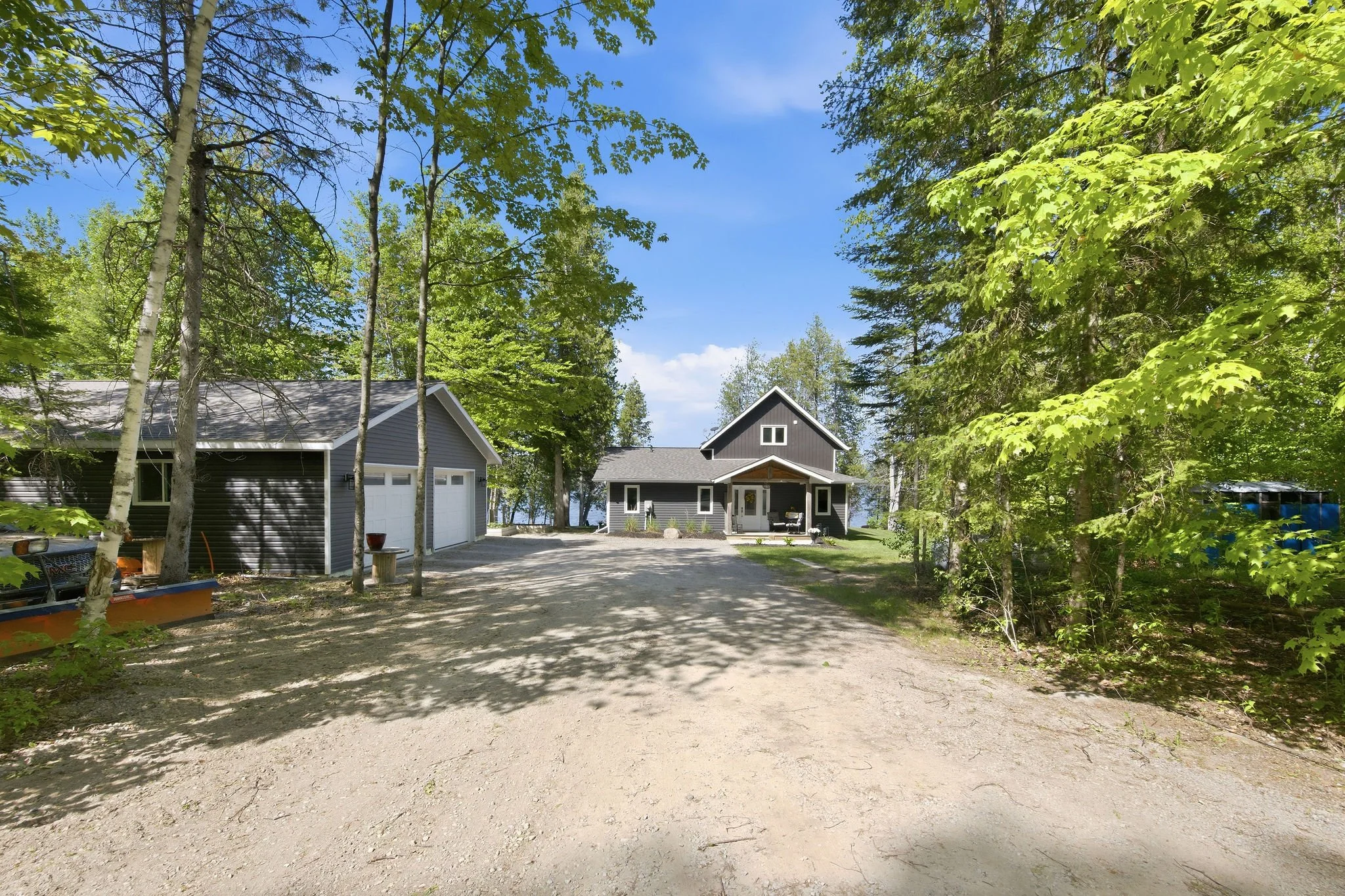 A gravel driveway leading to a house surrounded by tall trees and greenery. The house has dark gray siding and a front porch with outdoor furniture. A separate garage is nearby, and the scene is bright under a clear blue sky.