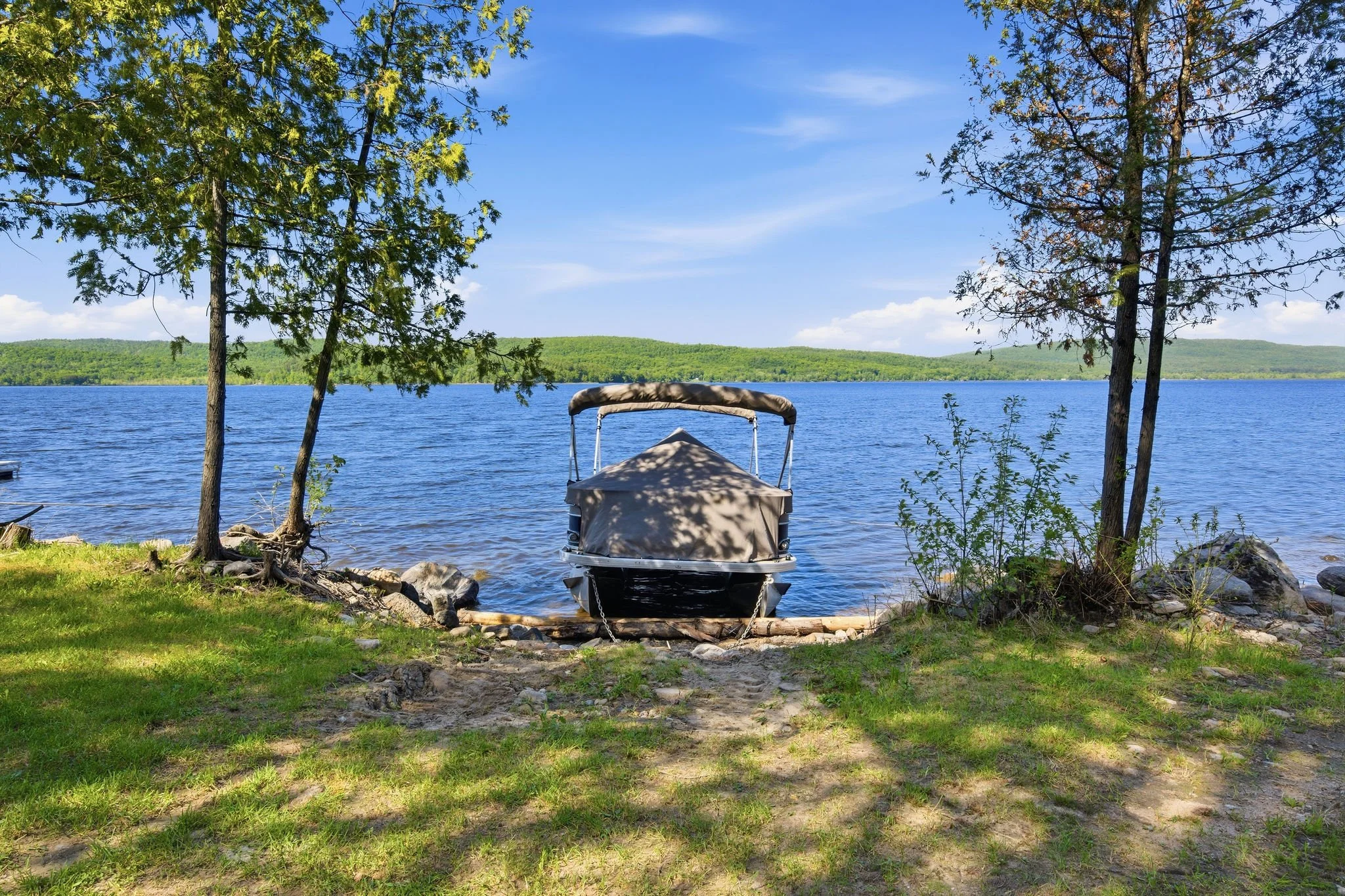 A boat docked on the shore of a lake with trees on each side and a green hill in the background under a blue sky with clouds.