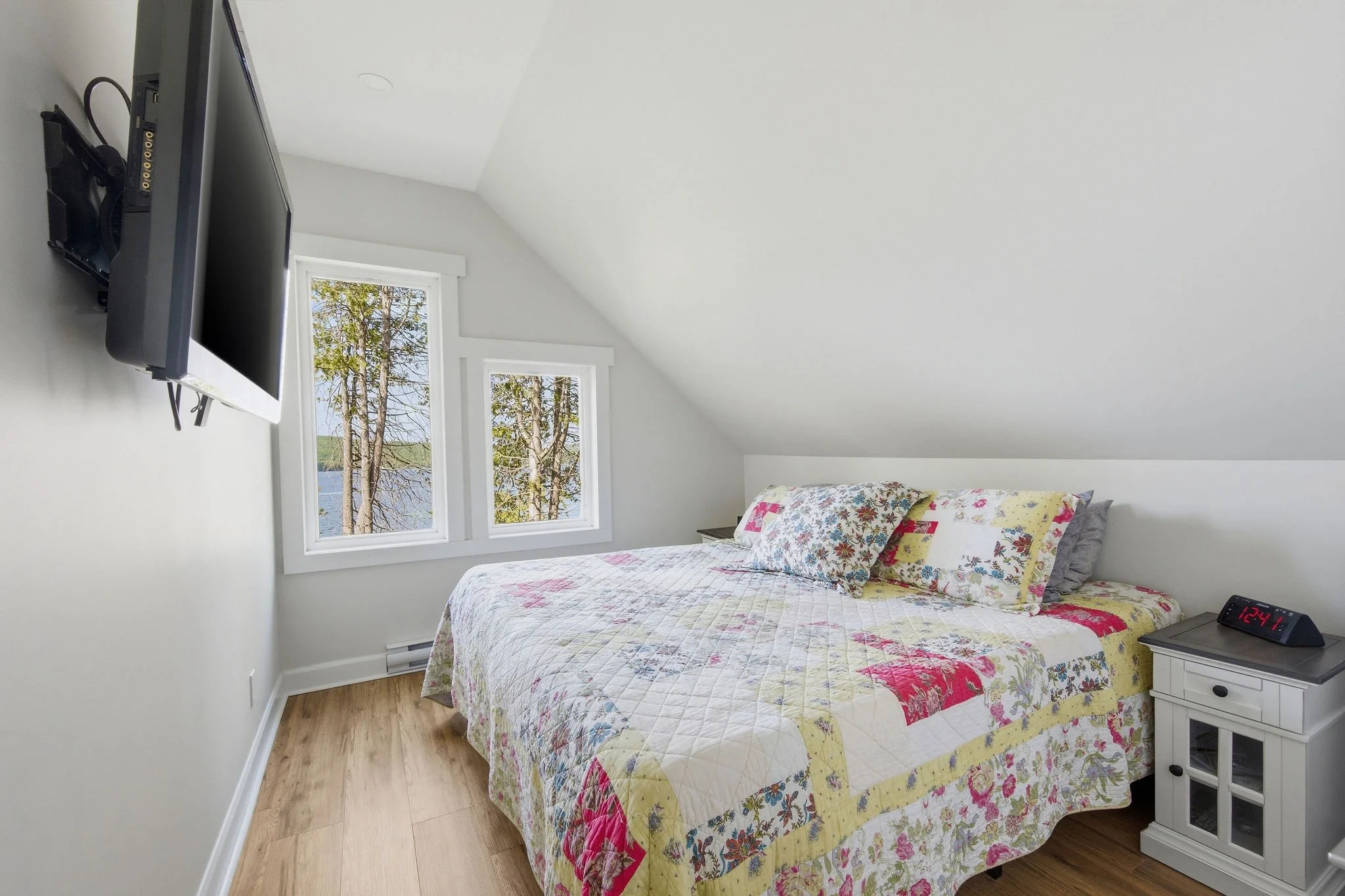 A bedroom with a bed featuring a colorful floral quilt and pillows, white nightstand with a clock showing 12:41, wall-mounted TV, and two windows with trees and a lake view.