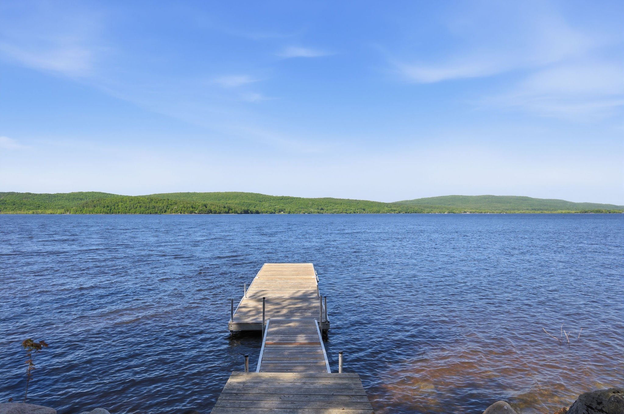 A wooden dock extends into a large blue lake, with green hills and a partly cloudy sky in the background.