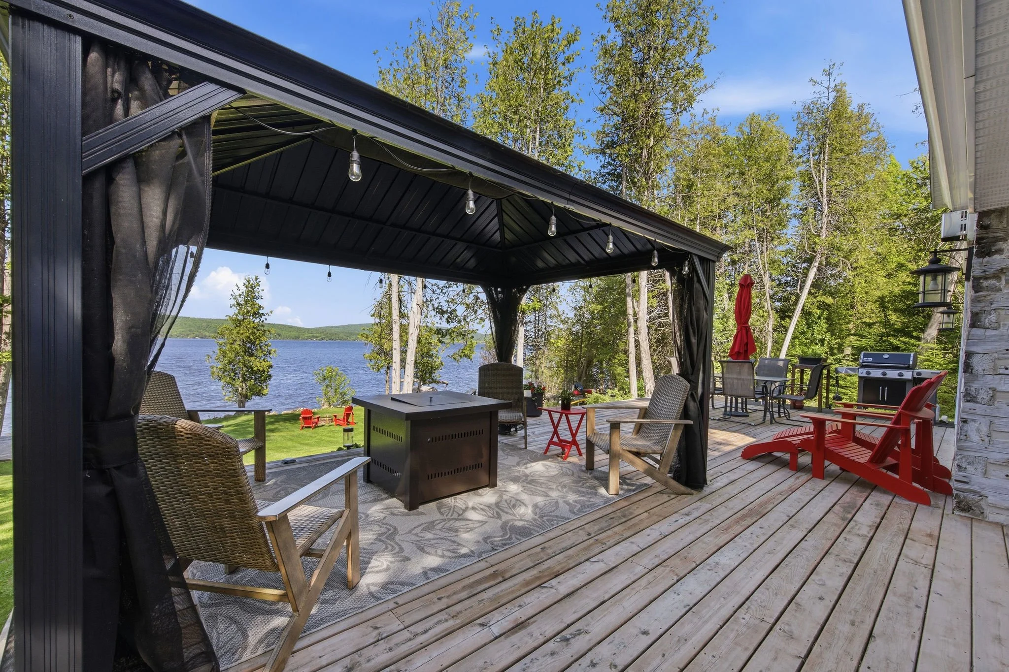 A covered outdoor patio overlooking a lake with trees in the background, featuring outdoor furniture including chairs, a grill, and string lights overhead.