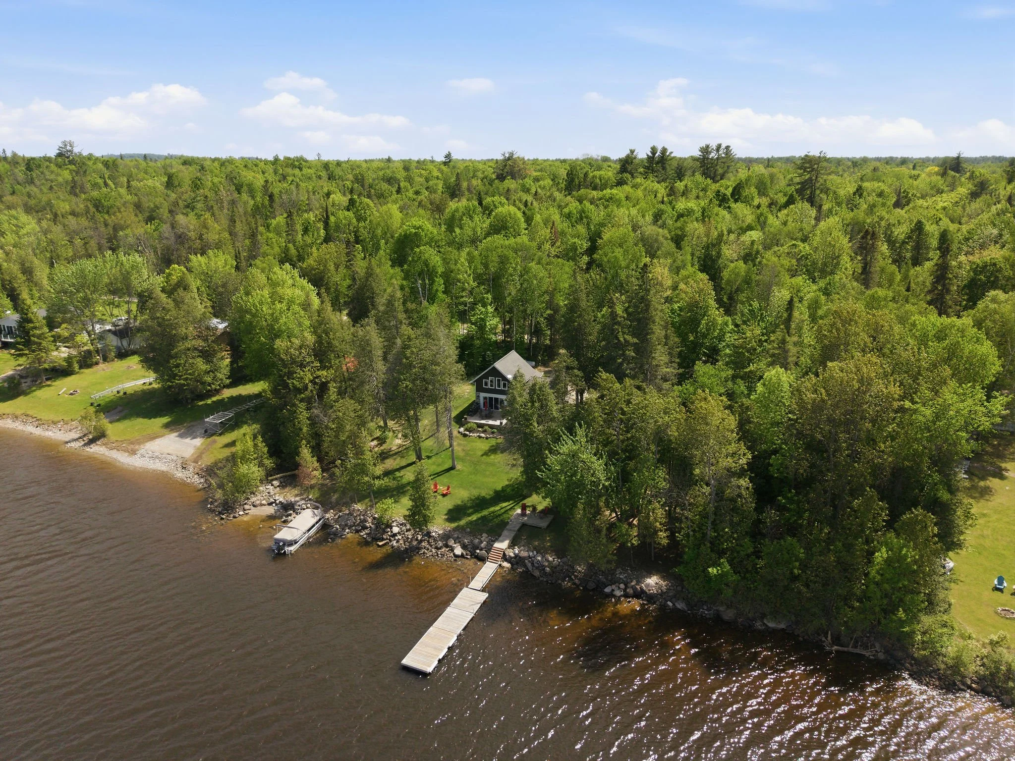Aerial view of a lakeshore with a small house, boats, and a dock surrounded by dense green trees on a sunny day.